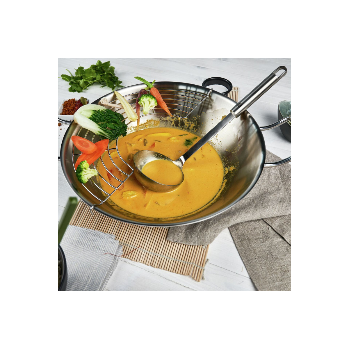 Wok z żółtym curry, warzywami i chochlą. Składniki z boku, na białej powierzchni.