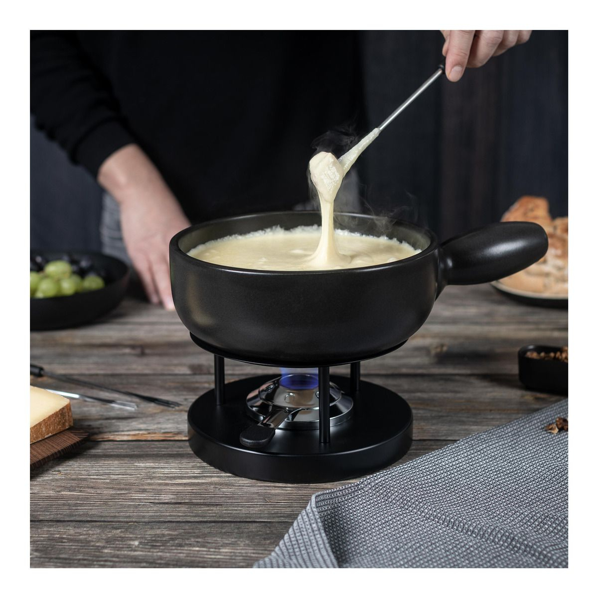 Czarny garnek do fondue z wyjmowanym serem, drewniany stół i szara serwetka.