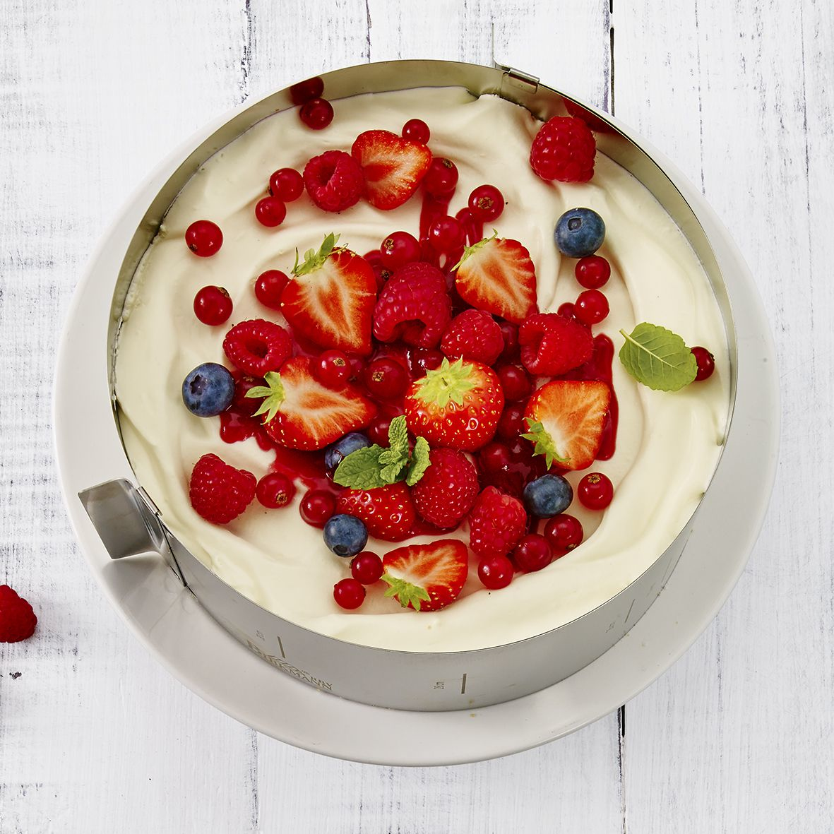 Kuchen mit Schlagsahne und Beeren in einem Ring auf weißer Holzoberfläche.