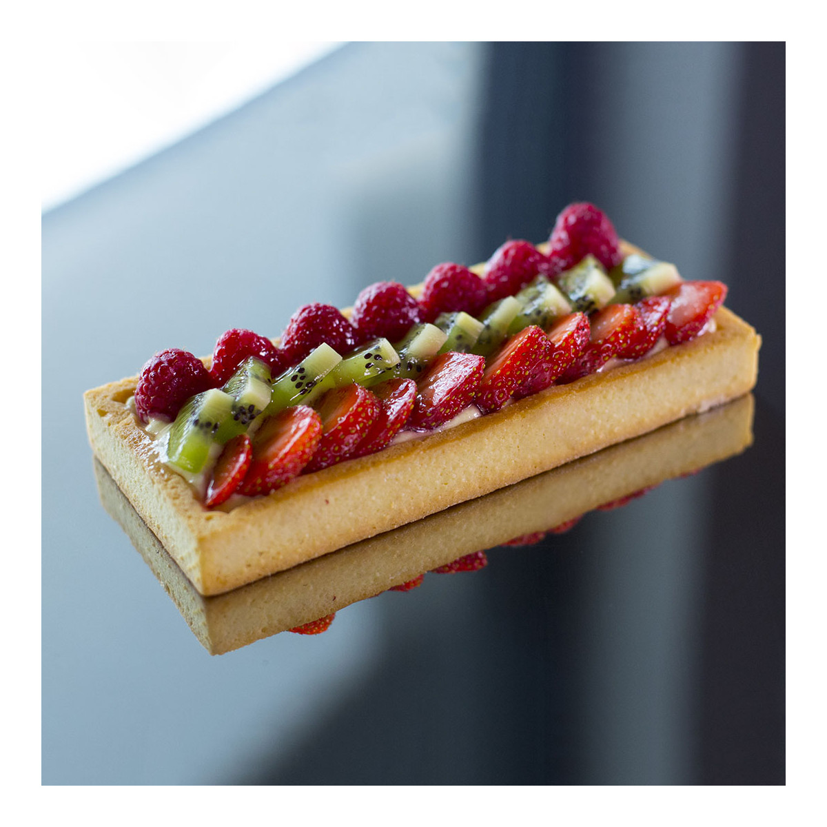 Ein rechteckiges Dessert mit Beeren und Kiwi, spiegelt sich auf glänzender Oberfläche.