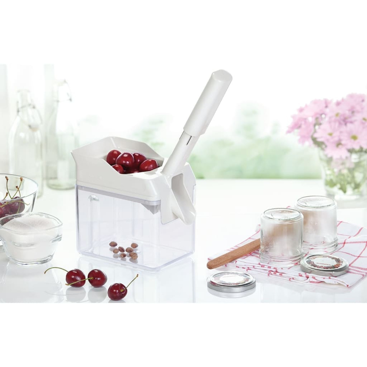 Biały drylownica do wiśni z wiśniami. Cukier, słoiki i kwiaty w tle.