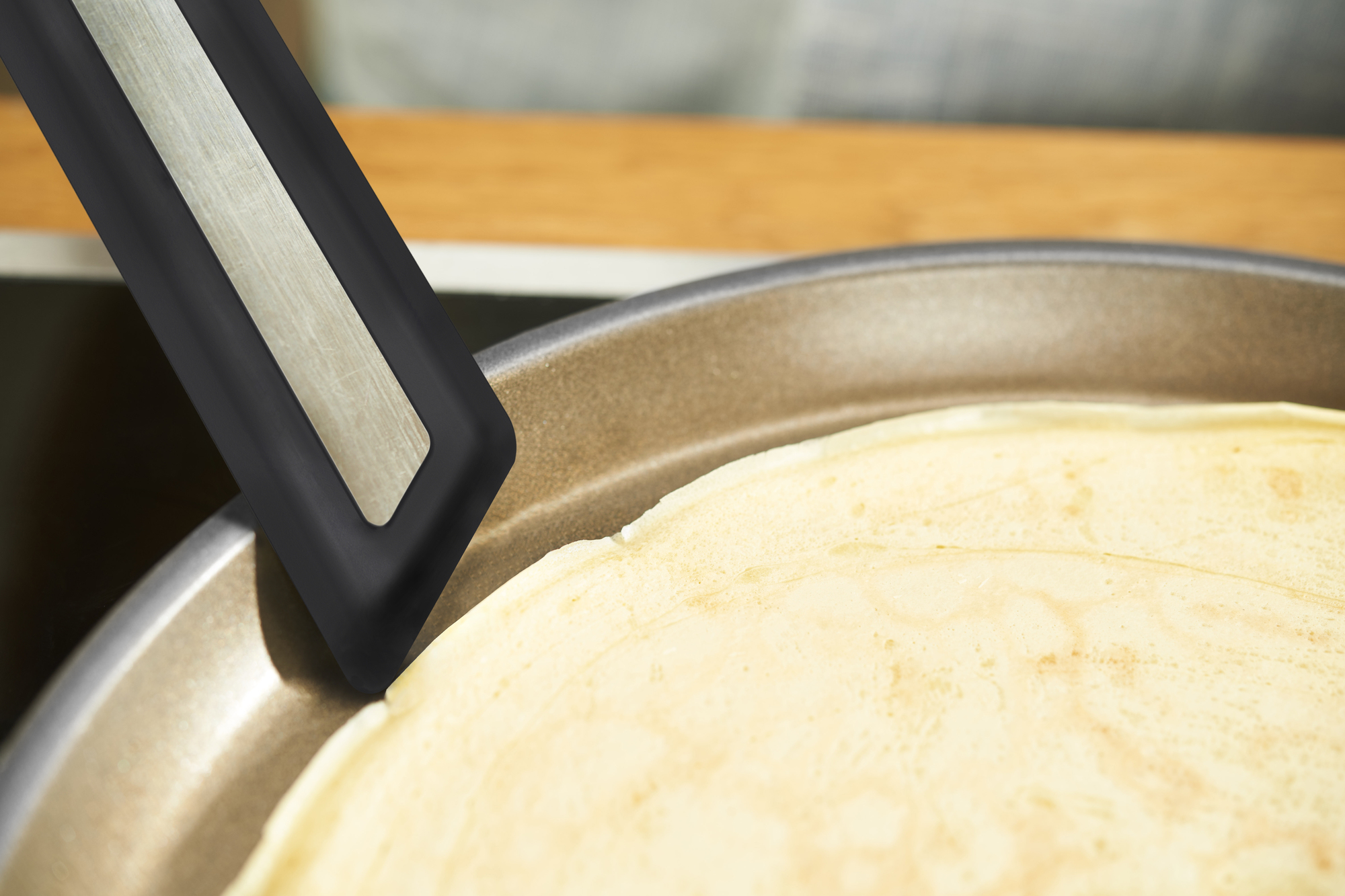 Ein Pfannenwender dreht einen Crêpe in einer Pfanne. Die Pfanne befindet sich auf weißem Hintergrund.