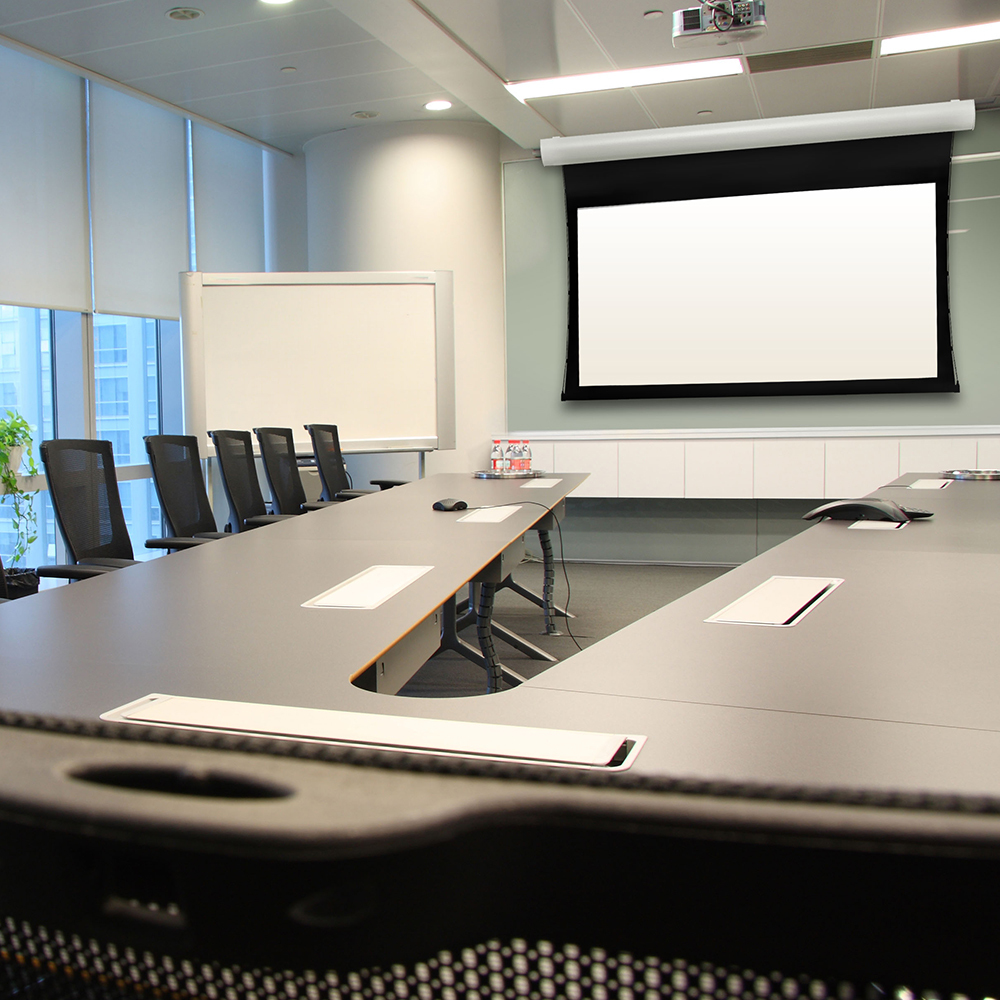 Sala conferenze con tavolo a forma di U. Schermo proiettore sullo sfondo. Sedie e una lavagna sono presenti.