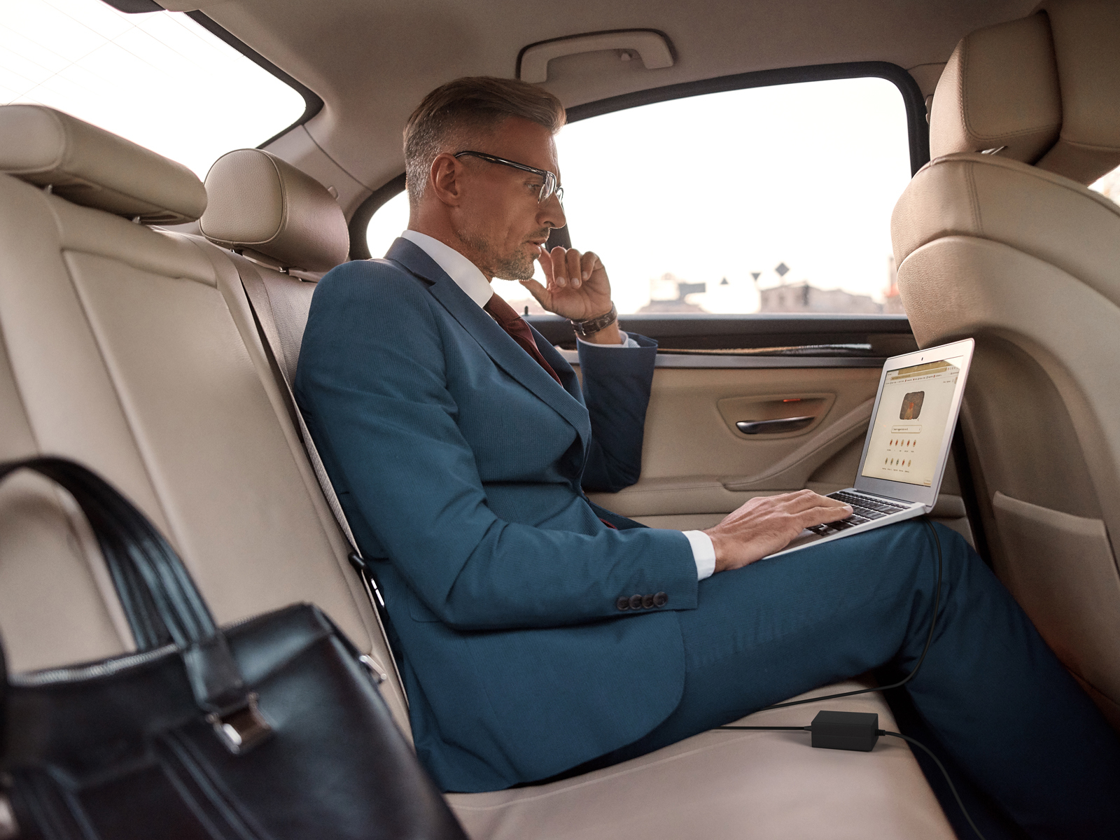 Mężczyzna w garniturze używa laptopa w samochodzie. Czarna torba na pierwszym planie. Czarna ładowarka.