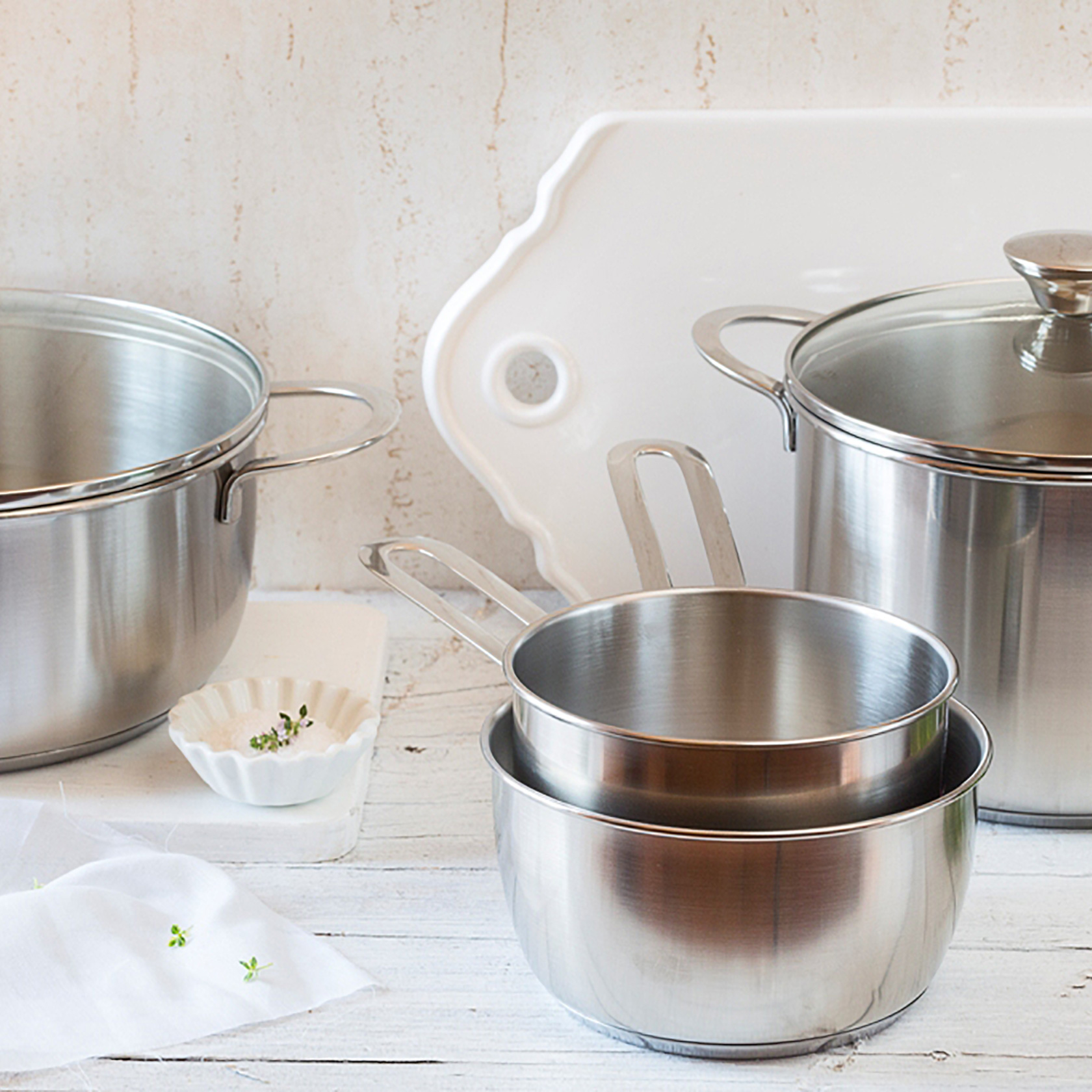 Kilka garnków, misek i deska do krojenia ze stali nierdzewnej na białej drewnianej powierzchni.