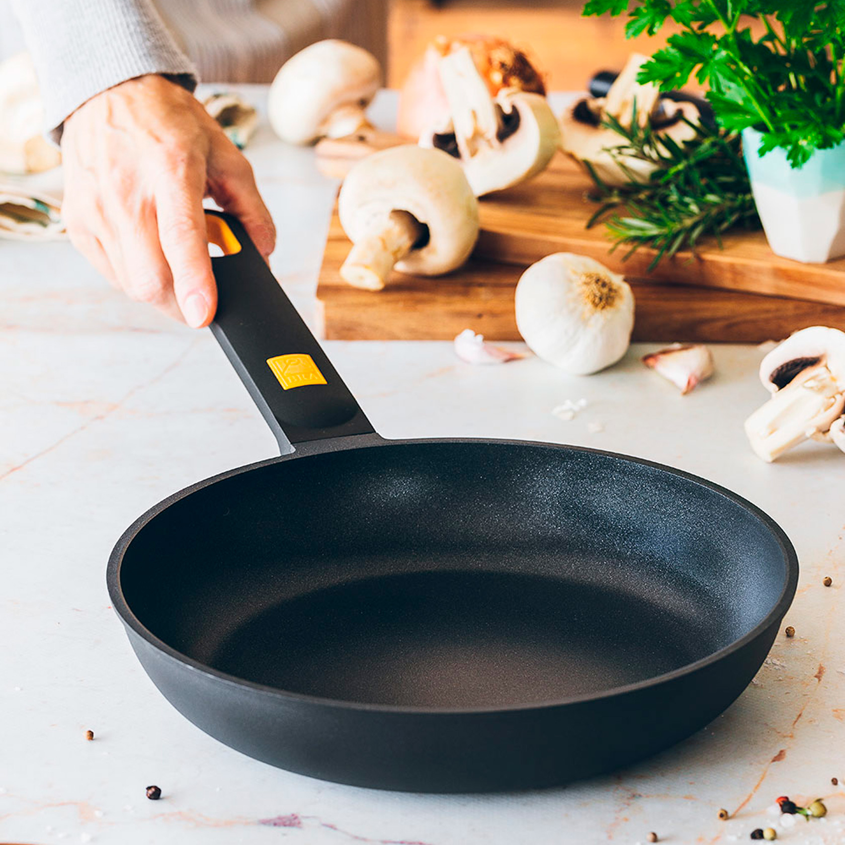 Dłoń trzymająca czarną patelnię z żółtym uchwytem, otoczoną składnikami.