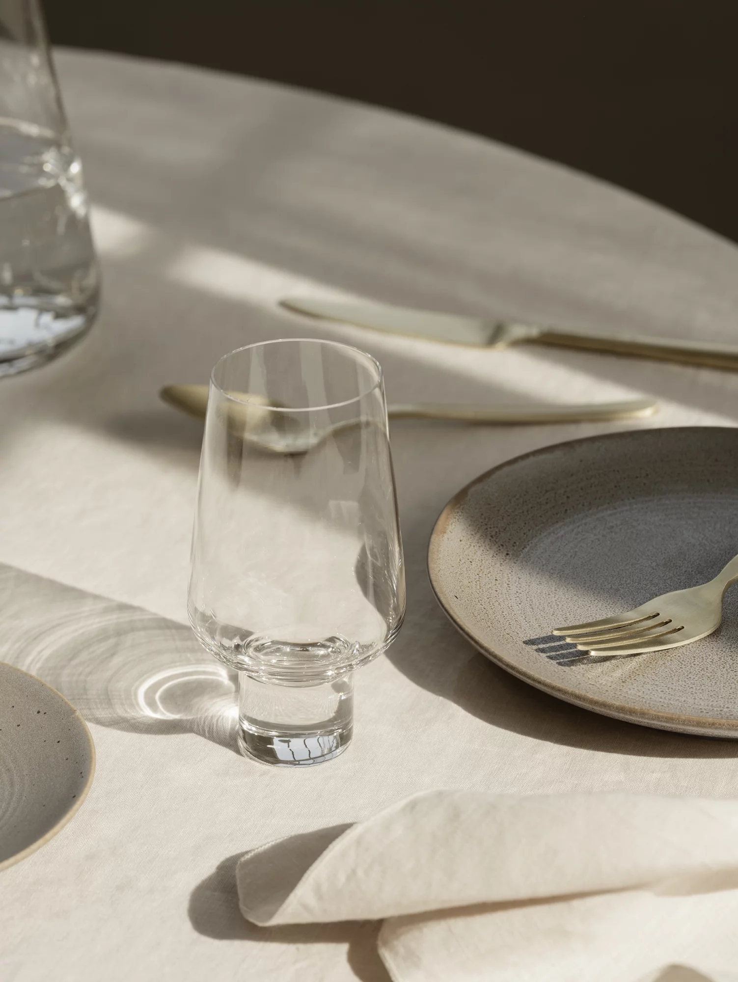 Ein hohes, klares Glas auf einem Tisch für ein Essen, mit einem Teller und Besteck. Weiches Sonnenlicht.