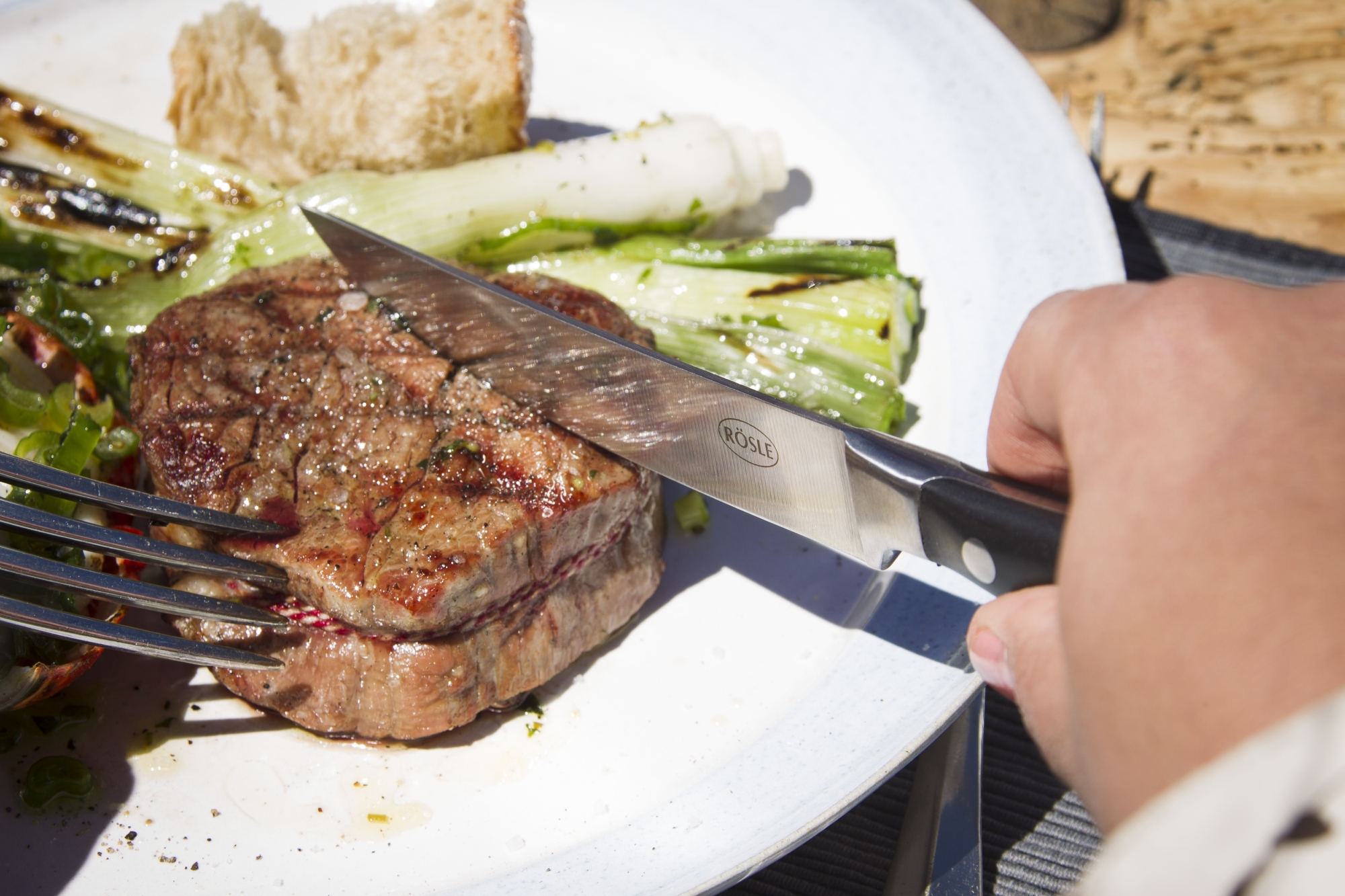 Eine Person schneidet ein gegrilltes Steak mit einem Messer auf einem weißen Teller mit gegrilltem Gemüse.