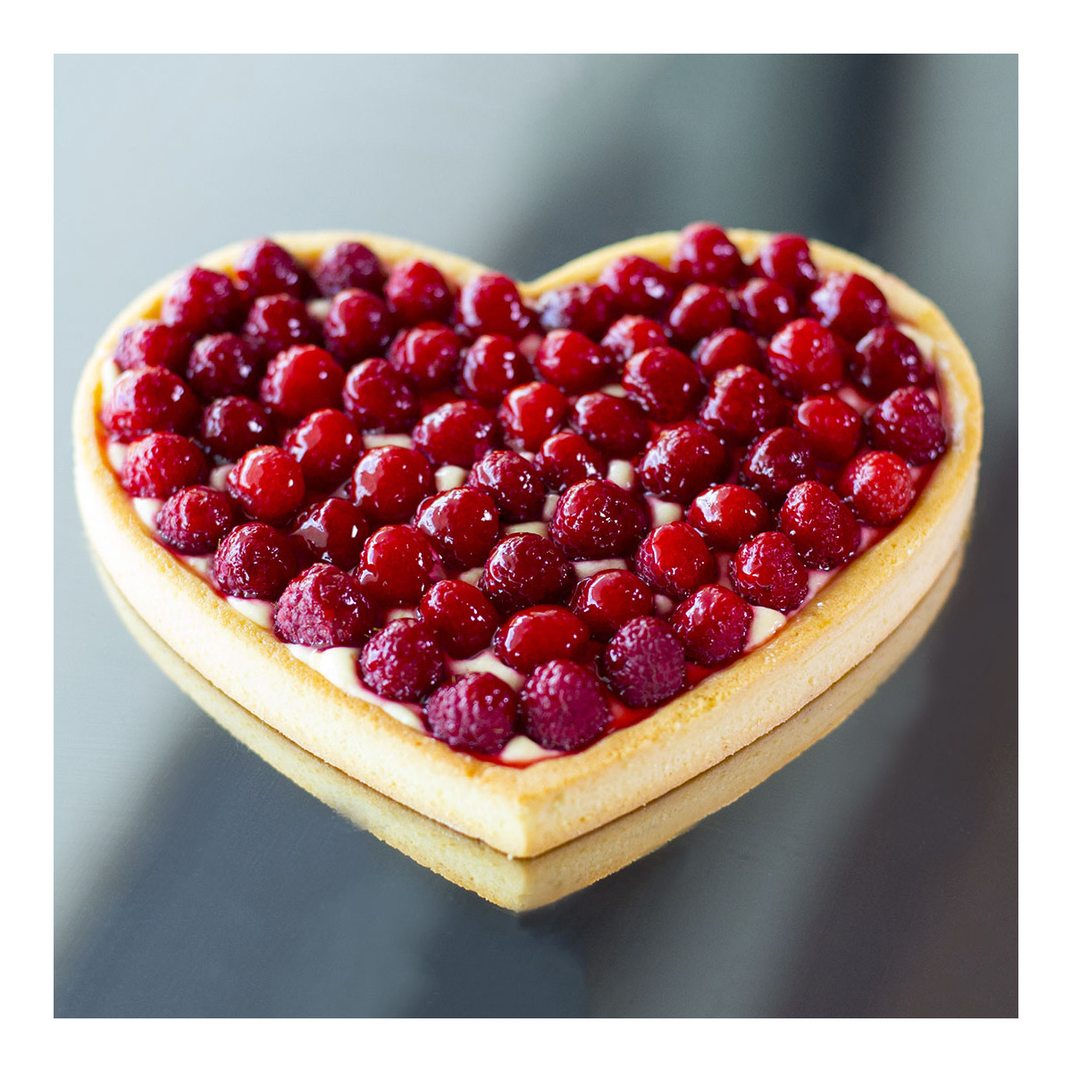 Eine herzförmige Tarte, belegt mit Himbeeren, auf einer verspiegelten Oberfläche.