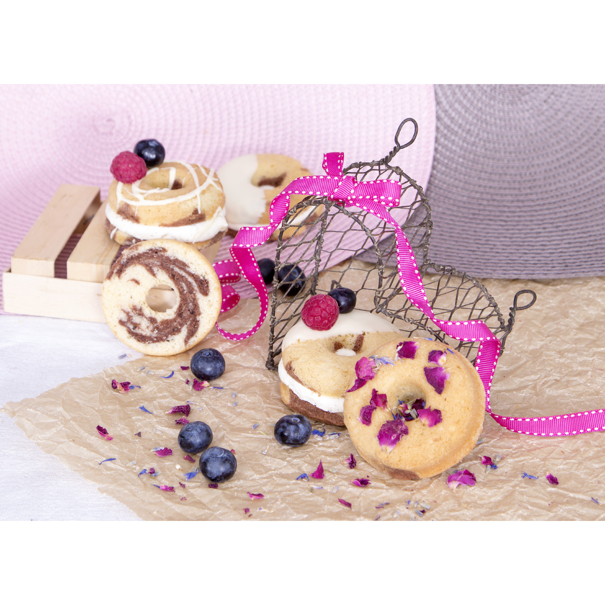 Donuts mit Sahne und Beeren auf einer Papieroberfläche. Ein rosa Band und Hüte sind im Hintergrund.