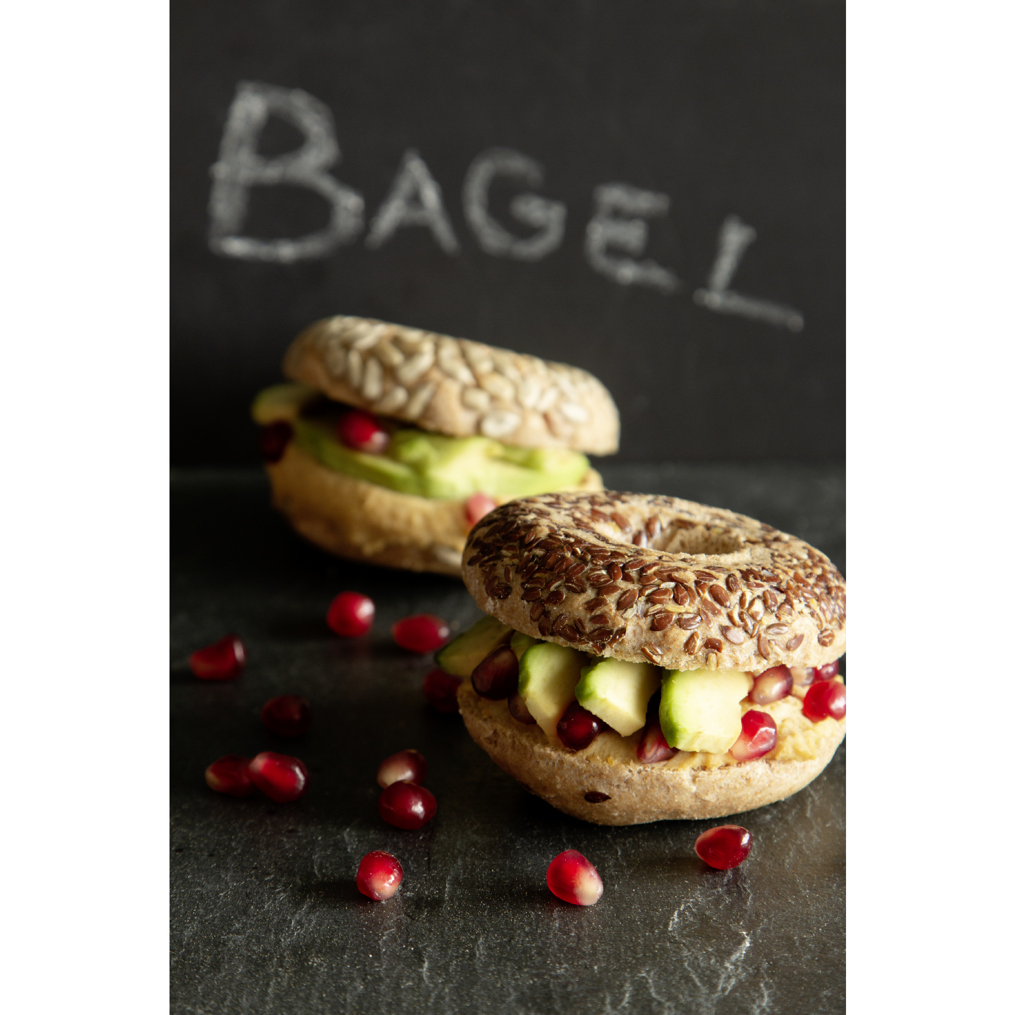 Zwei Bagels mit Avocado, Granatapfel und Grünzeug auf dunklem Schiefergrund.