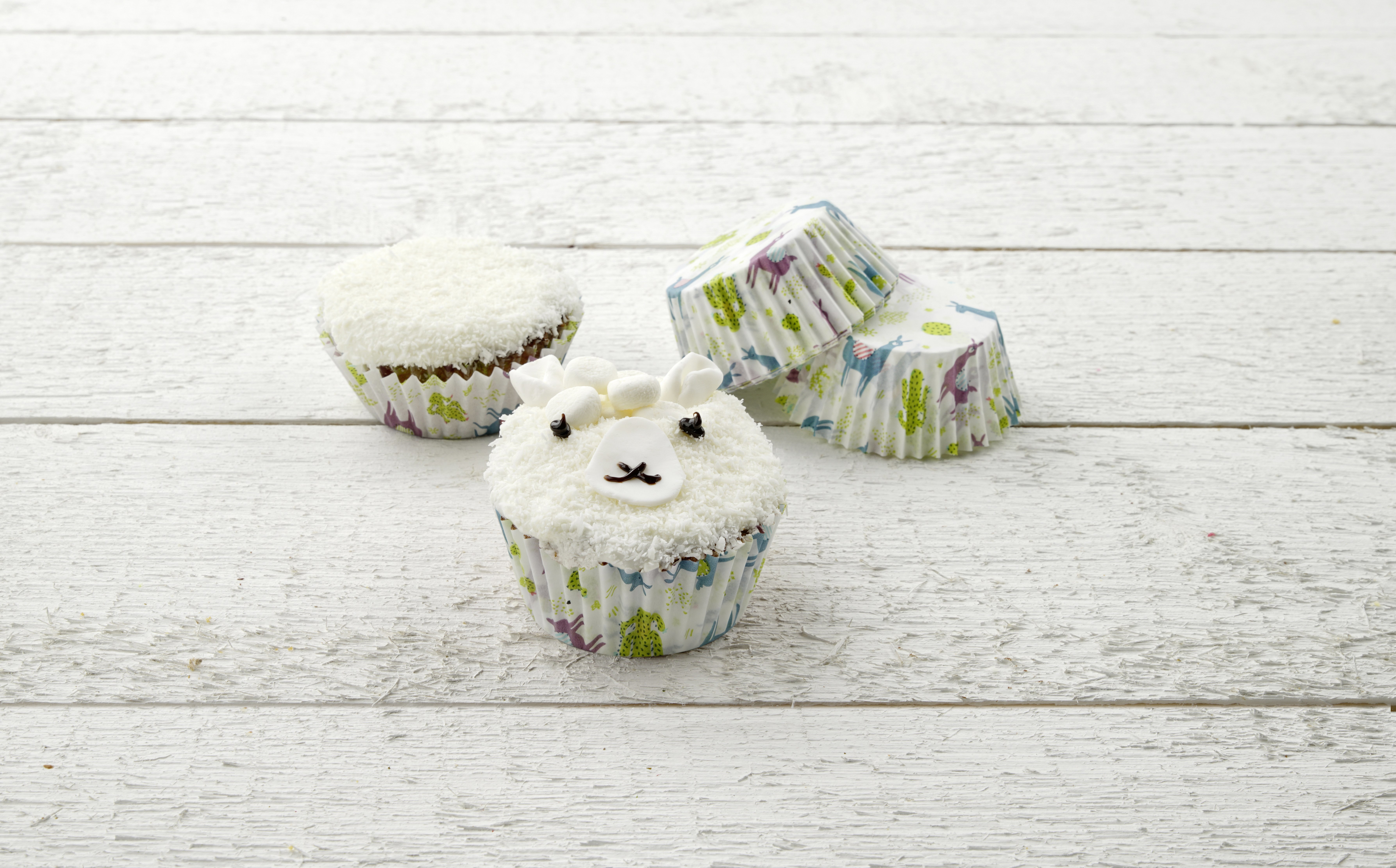 Drei Cupcakes mit Lama-Gesichtern auf einer weißen Holzoberfläche.