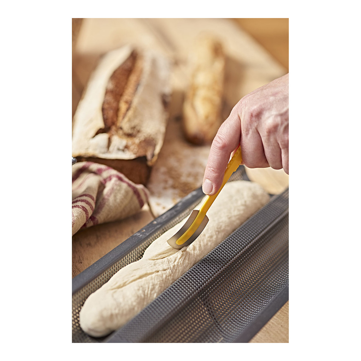 Eine Hand ritzt einen Brotlaib in einer Backform mit einer gelben Klinge ein.