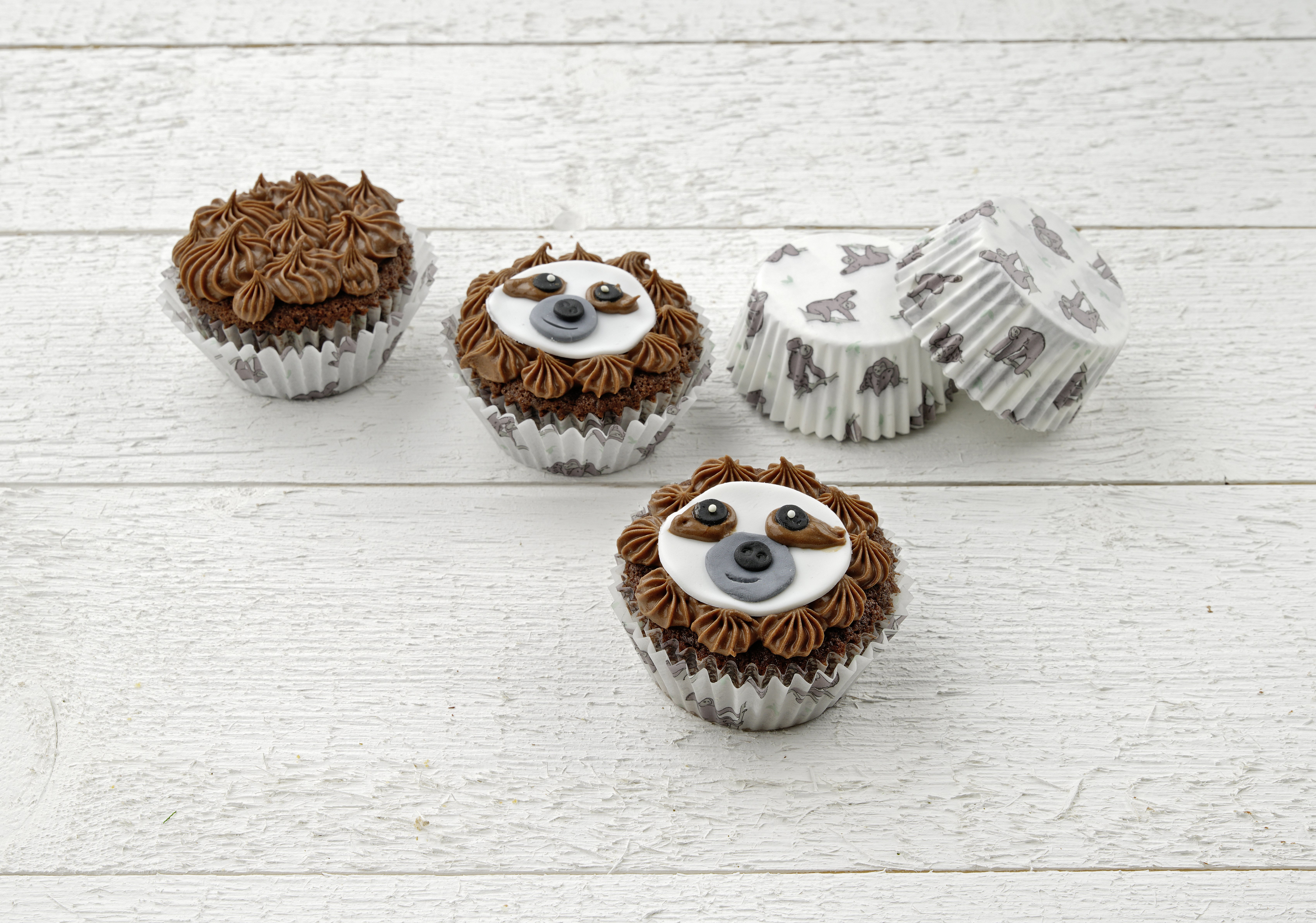 Drei Cupcakes mit braunem Zuckerguss und Faultiergesichtern, und zwei Cupcake-Formen mit Faultiermustern.
