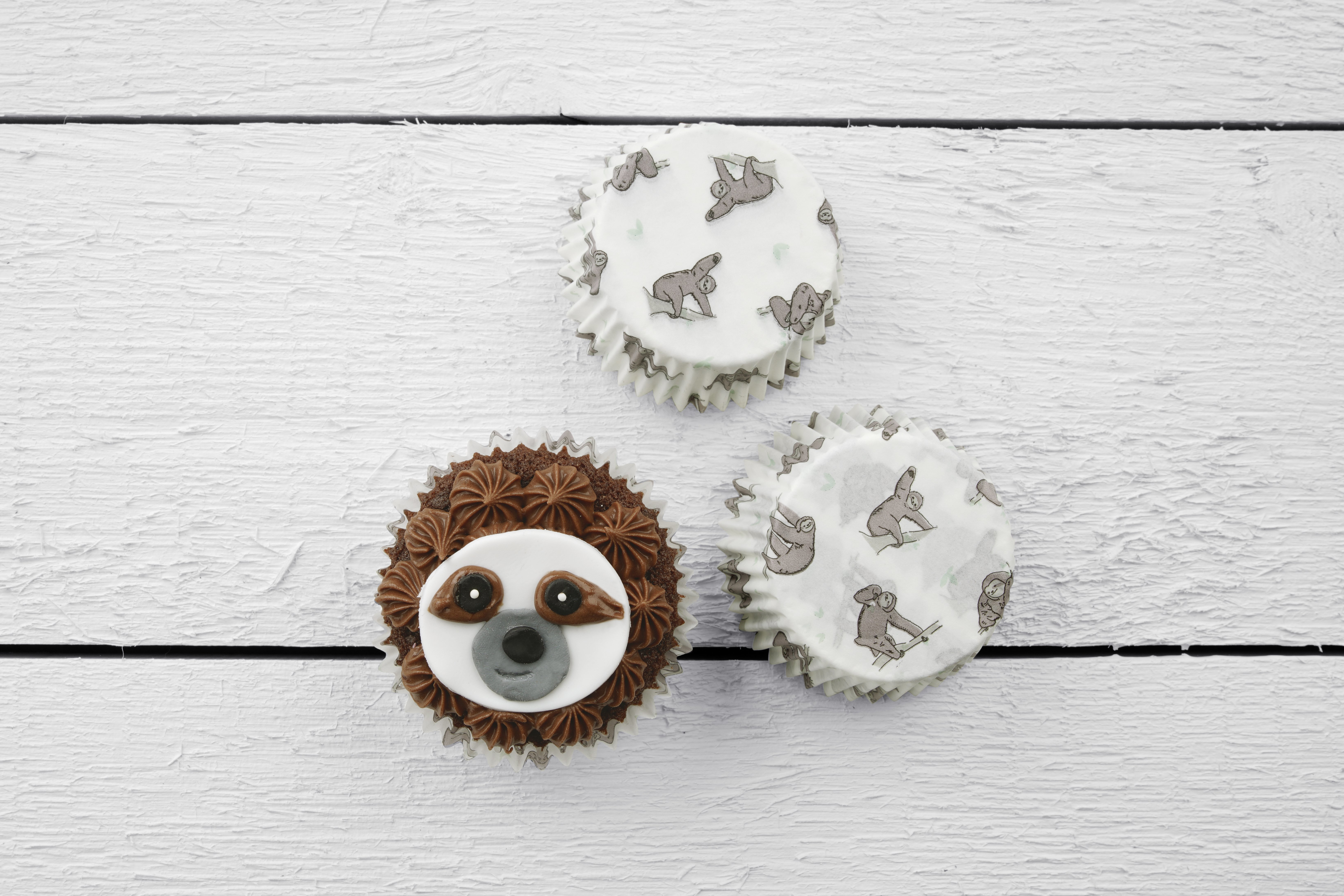 Drei Cupcakes, verziert mit braunem Zuckerguss, jeweils mit einem Faultiergesicht, auf weißem Brett.