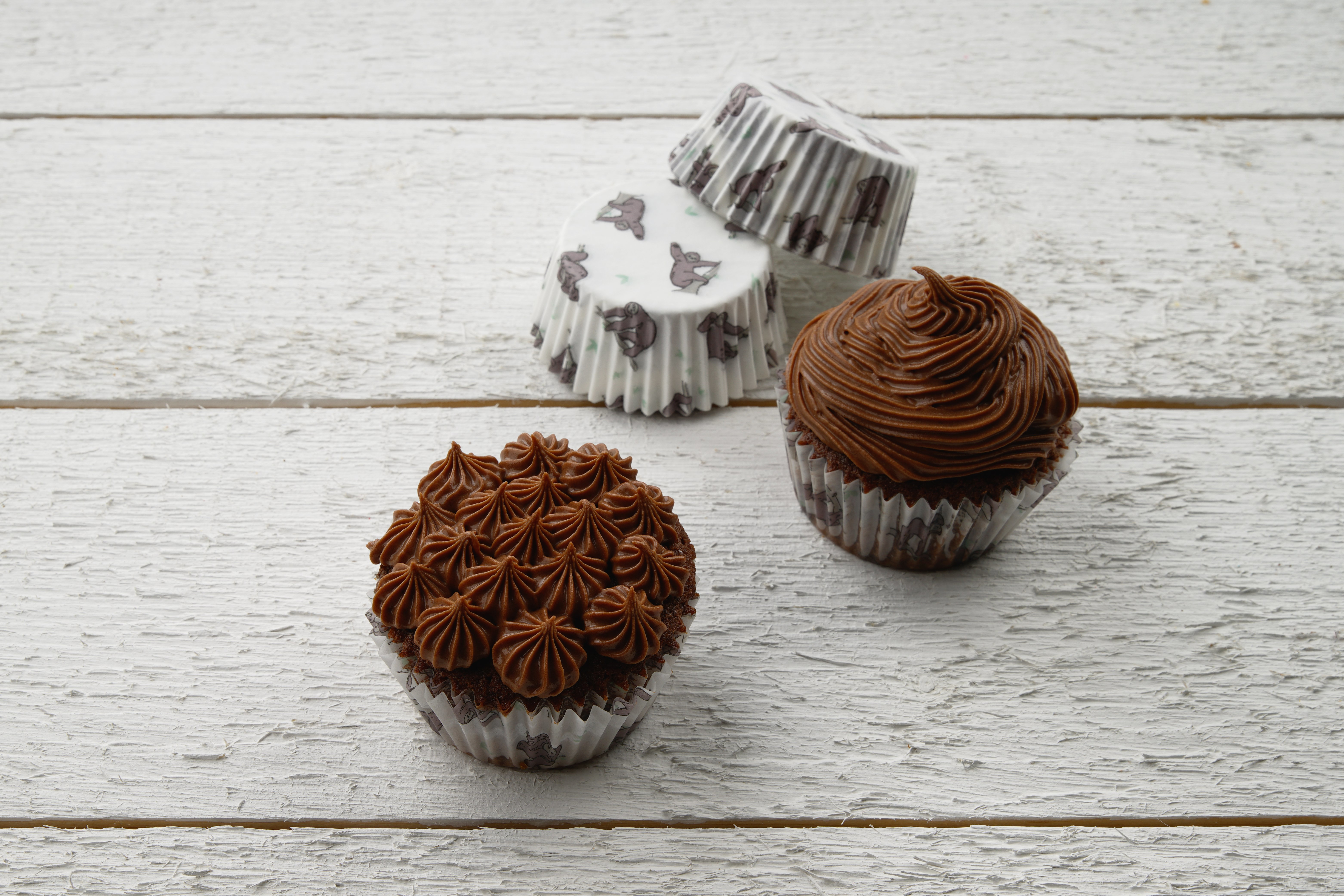 Zwei Cupcakes mit Schokoladenfrosting und einer mit Cupcake-Formen auf weißer Holzoberfläche.