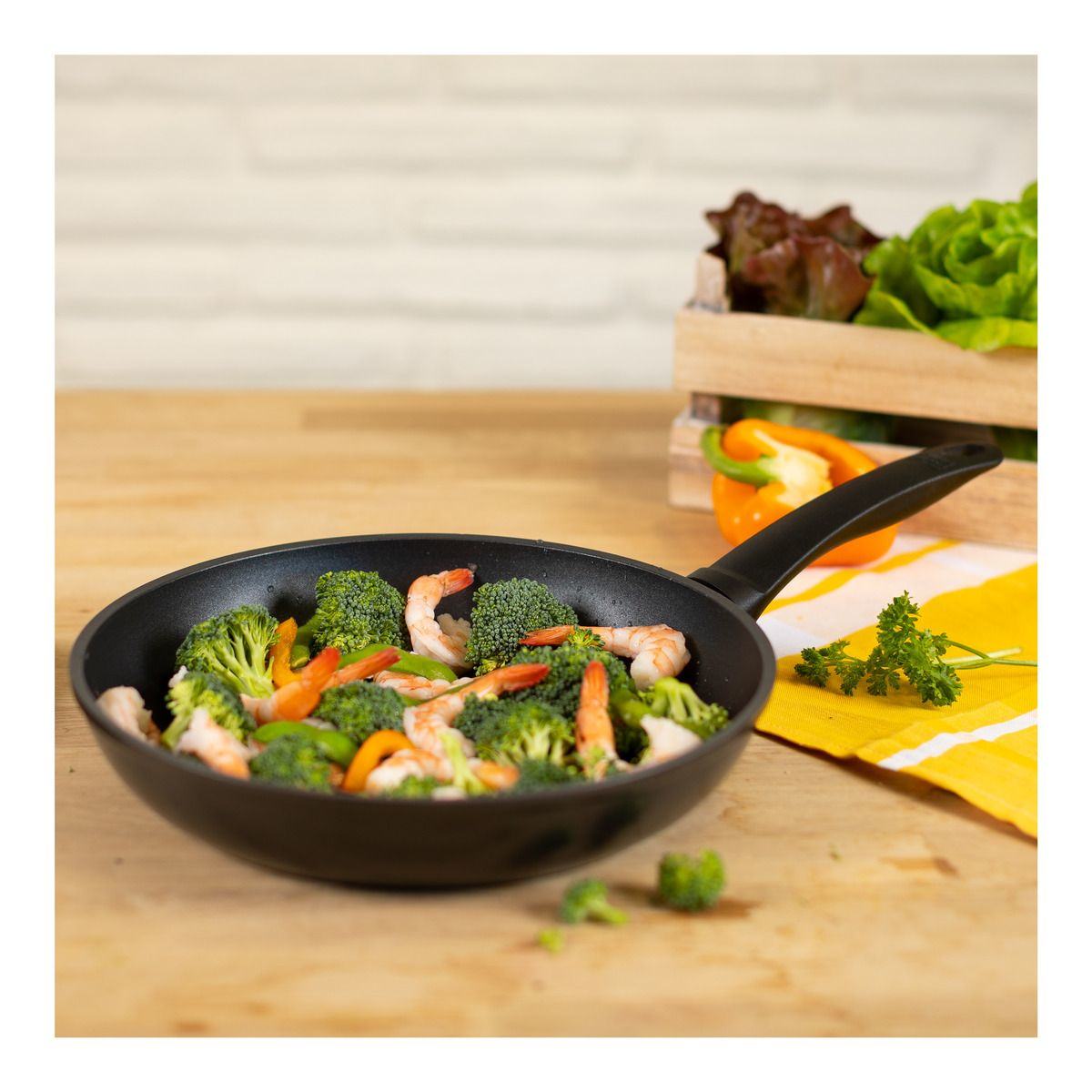 Sartén con camarones, brócoli y verduras; mesa de madera, lechuga y pimiento en el fondo.