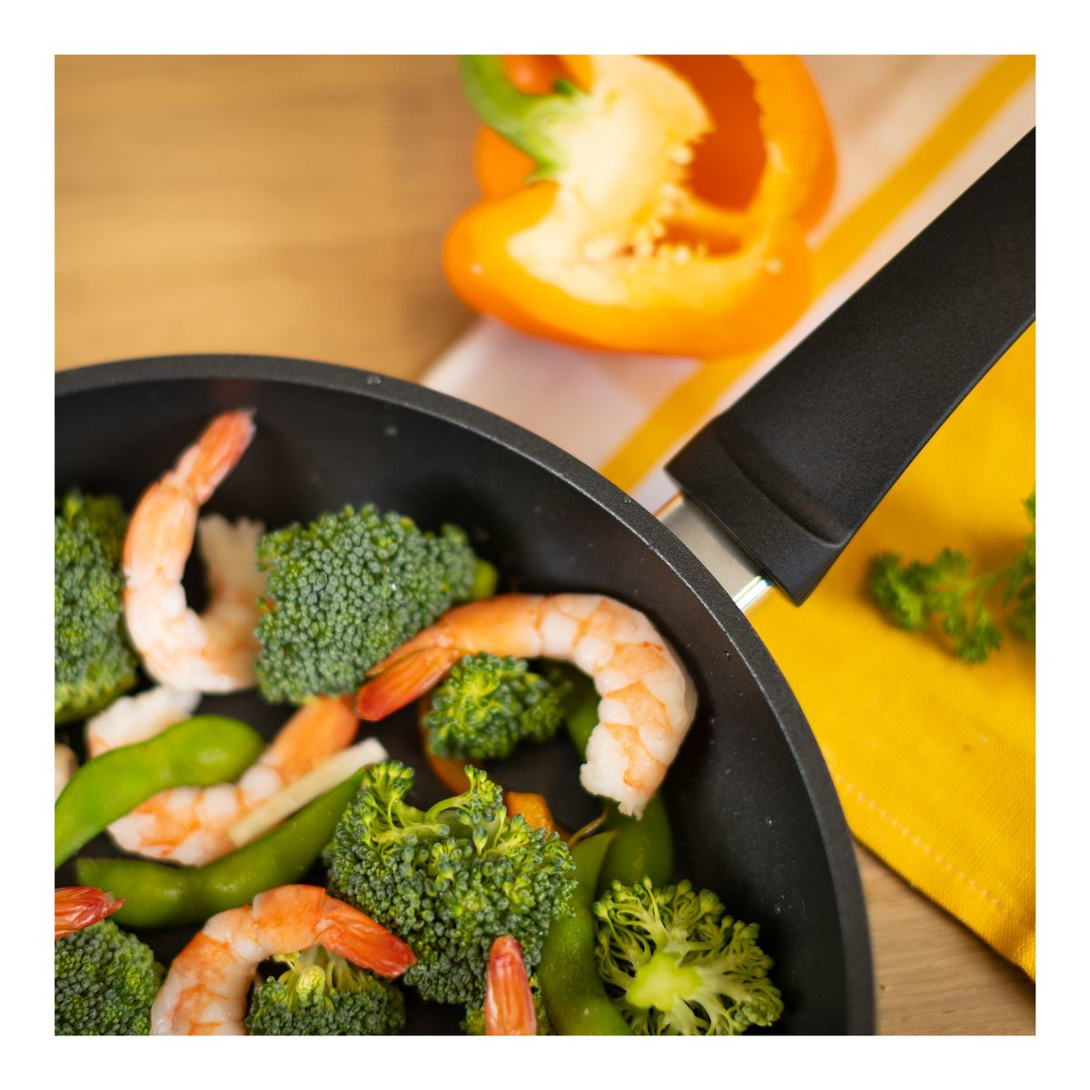 Sartén con camarones, brócoli y verduras; pimiento y servilleta en el fondo.