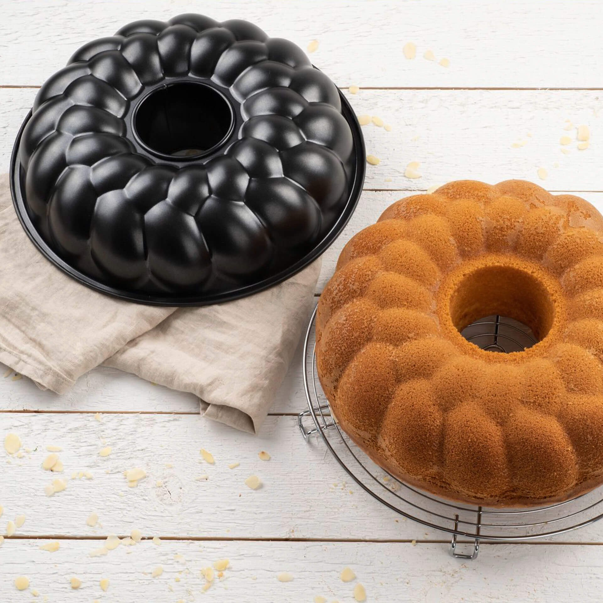 Eine schwarze Gugelhupf-Form und ein gebackener Kuchen mit Loch auf einer weißen Holzoberfläche mit Mandelblättchen.