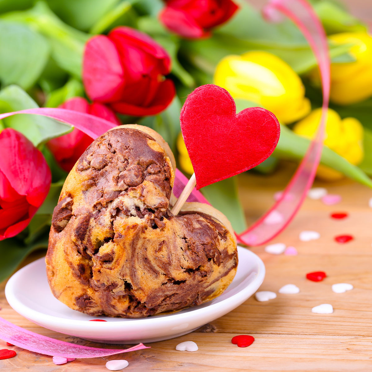 Ein herzförmiger Muffin mit rotem Herz und Tulpen im Hintergrund auf einer Holzoberfläche.