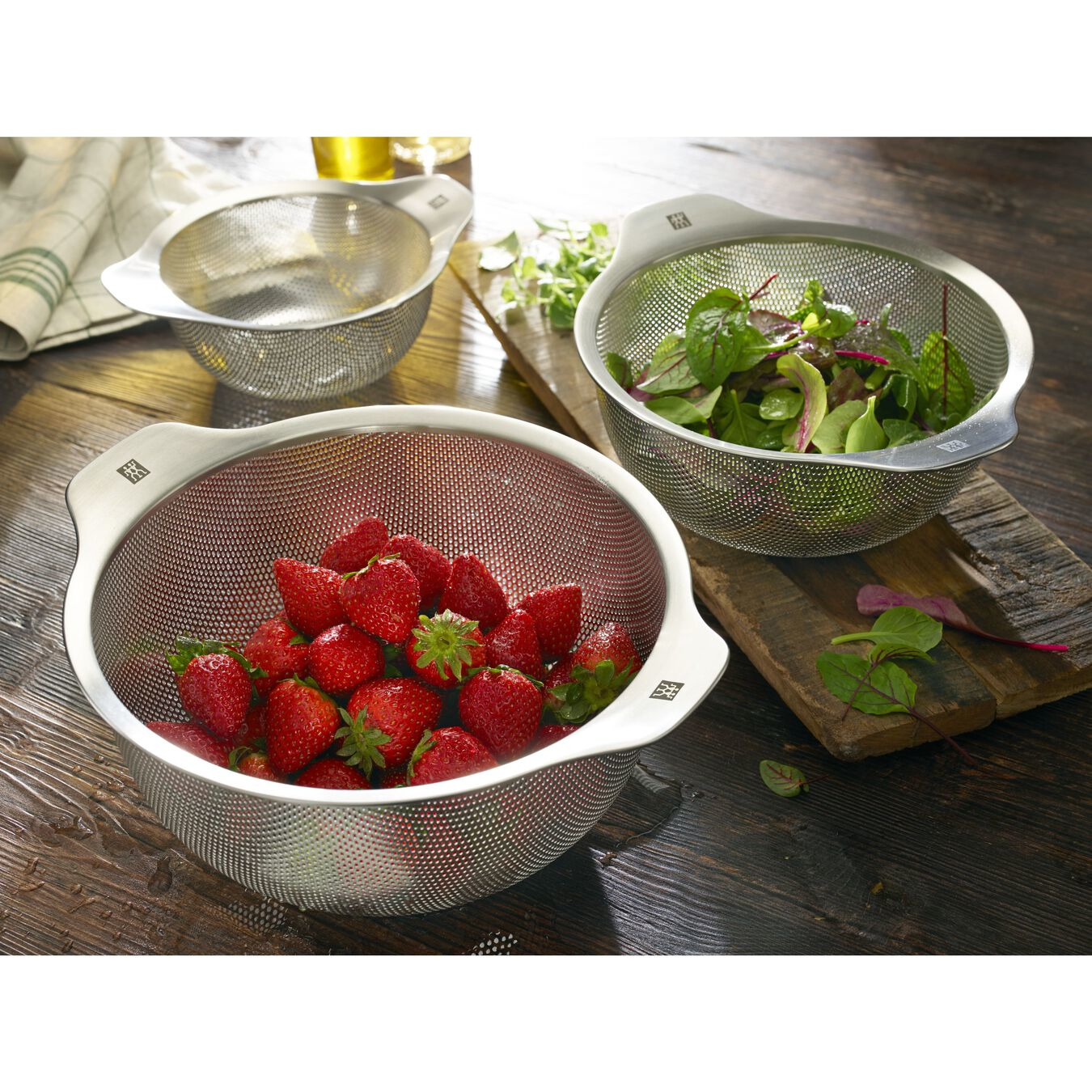 Three stainless steel colanders with strawberries and greens on a wooden surface.