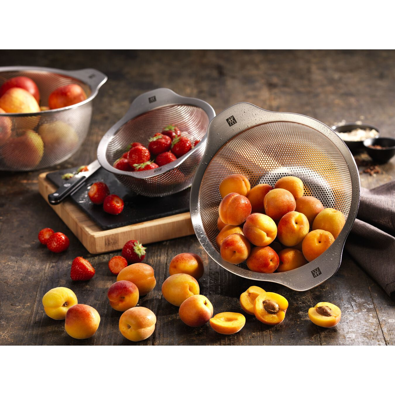 Stainless steel colanders with apricots and strawberries on a wooden surface.