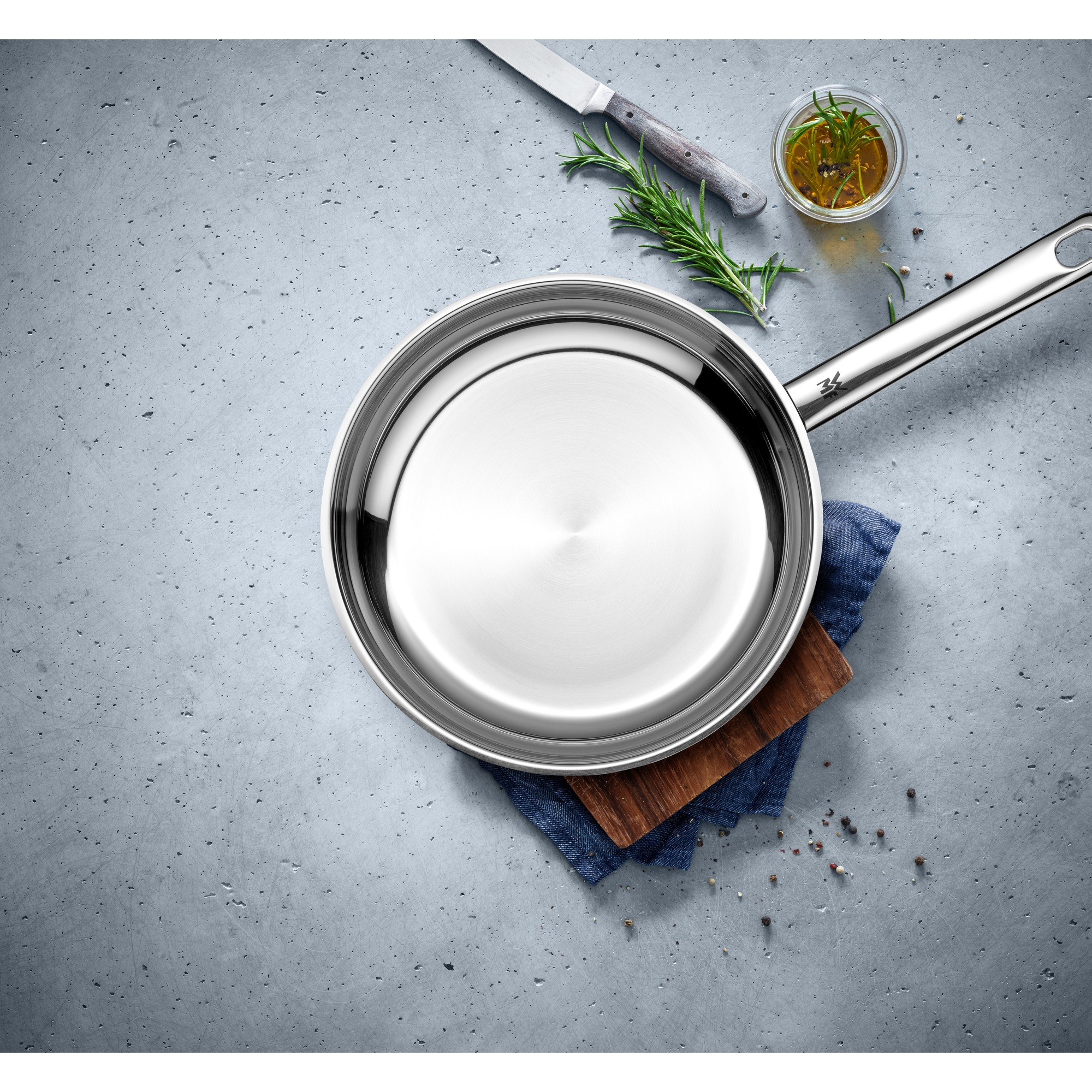 Patelnia z olejem i ziołami, umieszczona na niebieskiej szmatce na szarej powierzchni.