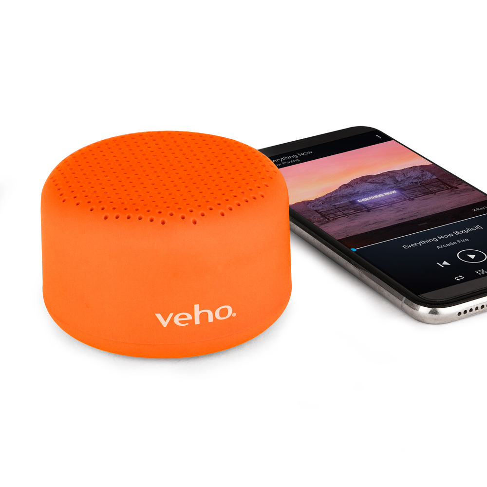 Altoparlante veho arancione accanto a un telefono che mostra un'app musicale, su una superficie bianca.