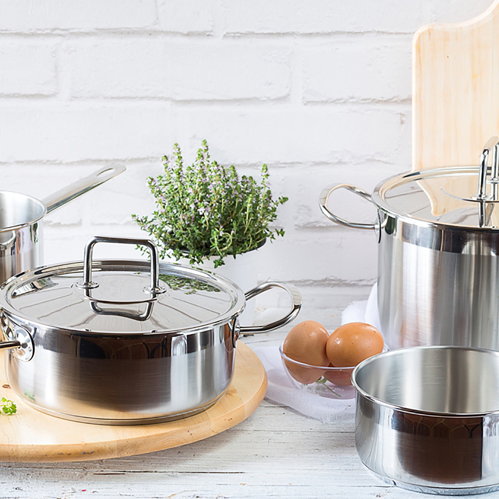 Garnki ze stali nierdzewnej z pokrywkami, jajka w misce i roślina na jasnej powierzchni.