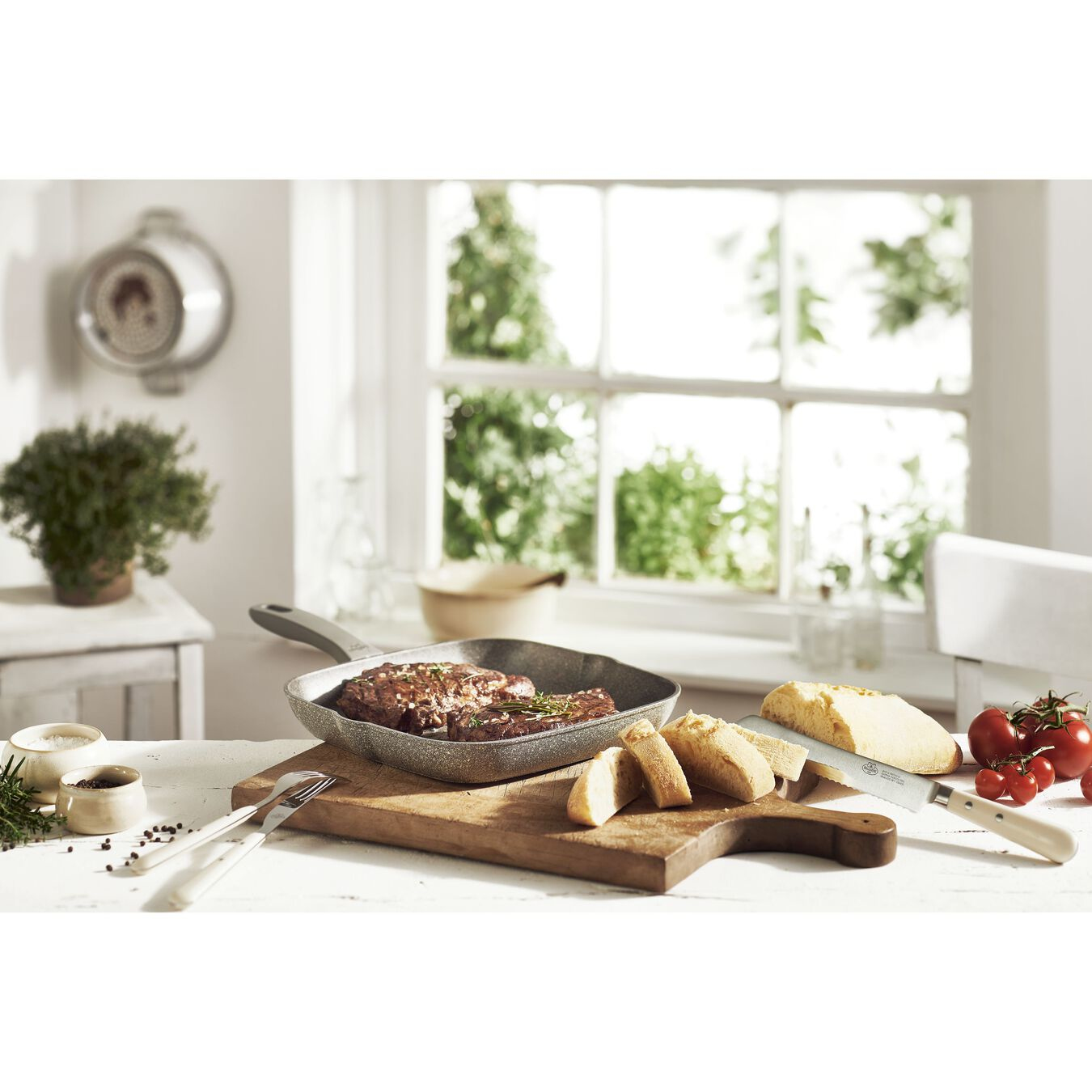 Küchenszene mit Grillpfanne, Steak, Brot und Gemüse auf einem Holzbrett.