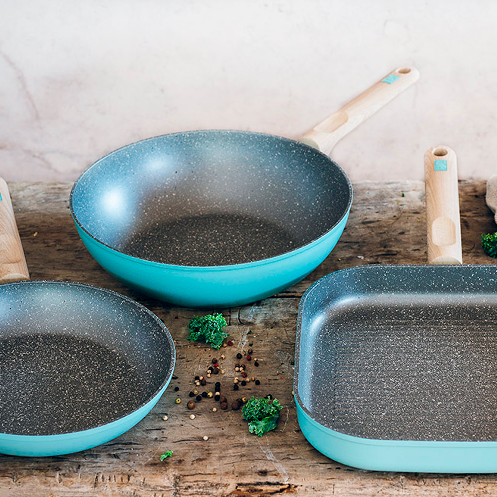 Trzy turkusowe patelnie na drewnianej powierzchni z przyprawami i zielenią. Patelnie mają nakrapiane szare wnętrze.