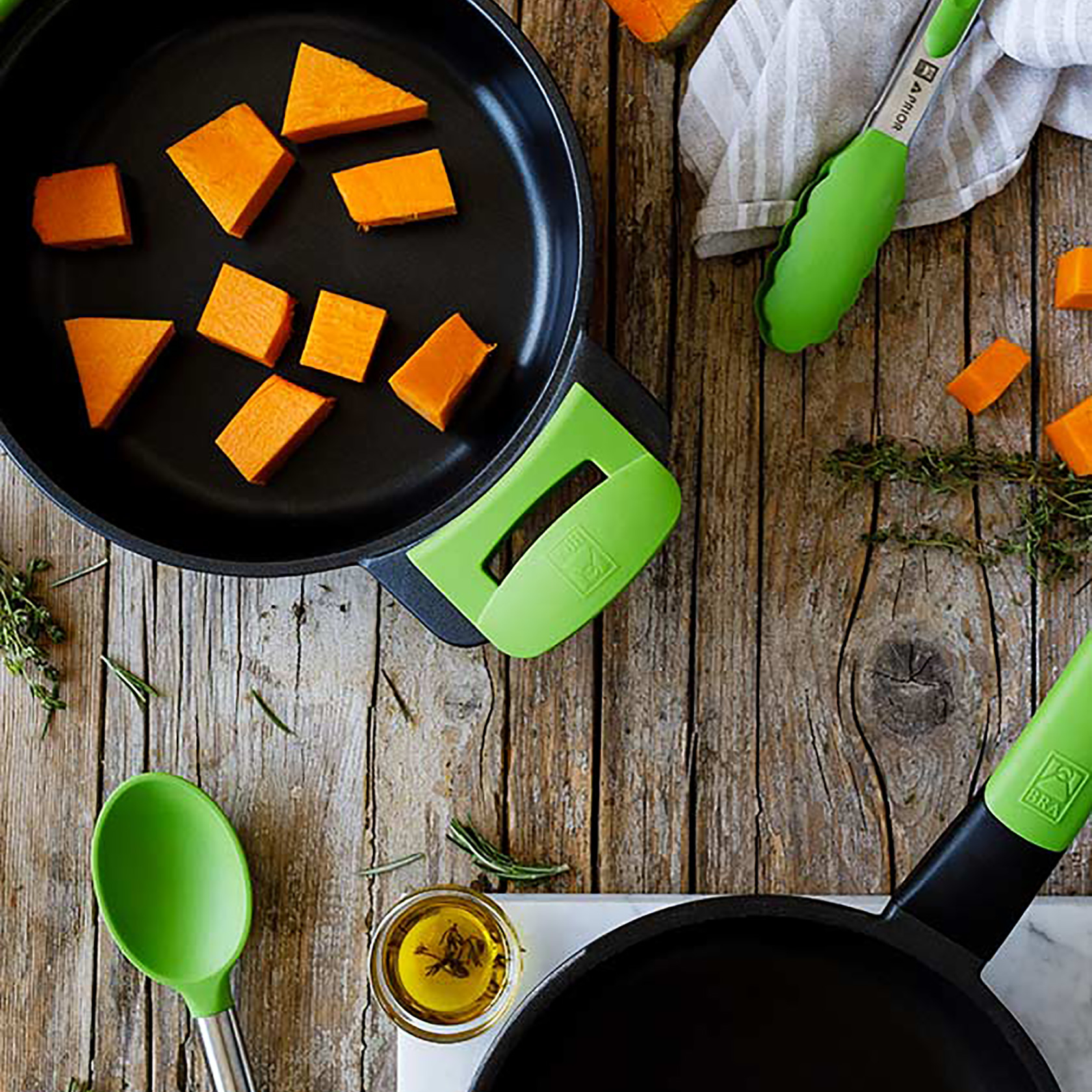 Widok z góry na drewniany stół z patelniami, pokrojoną dynią, zielonymi szczypcami i łyżką oraz olejem.