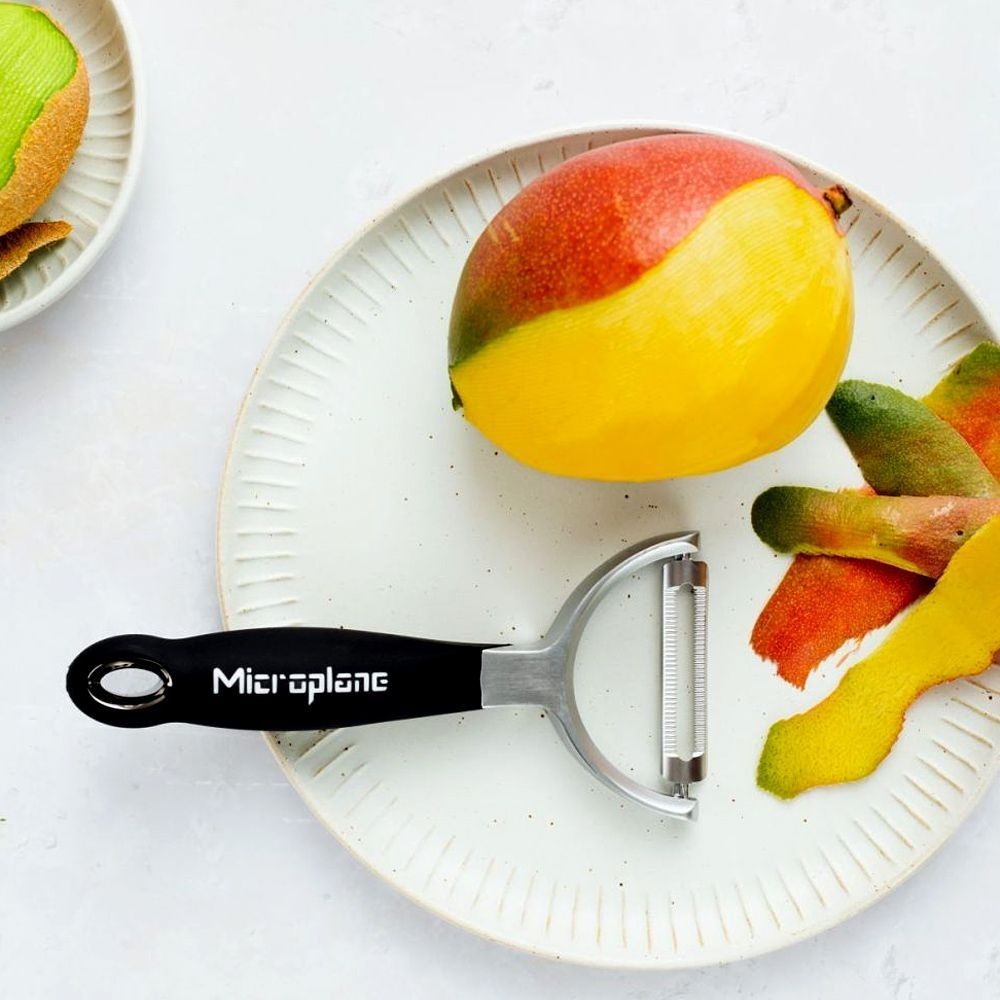 Mango auf einem weißen Teller mit einem schwarz-silbernen Schäler. Mangostücke sind ebenfalls vorhanden.