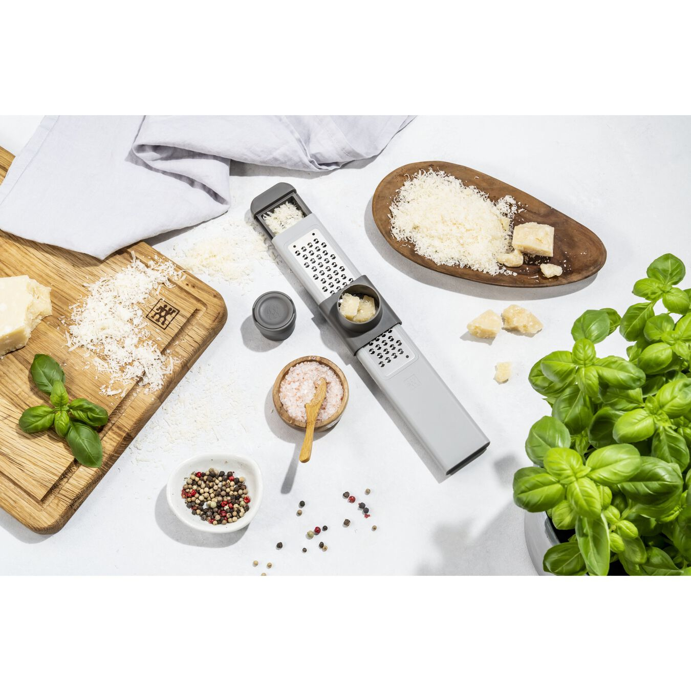 Kitchen scene with grater, basil, cheese, spices, and wooden boards. Bright, top-down view. Background: White.