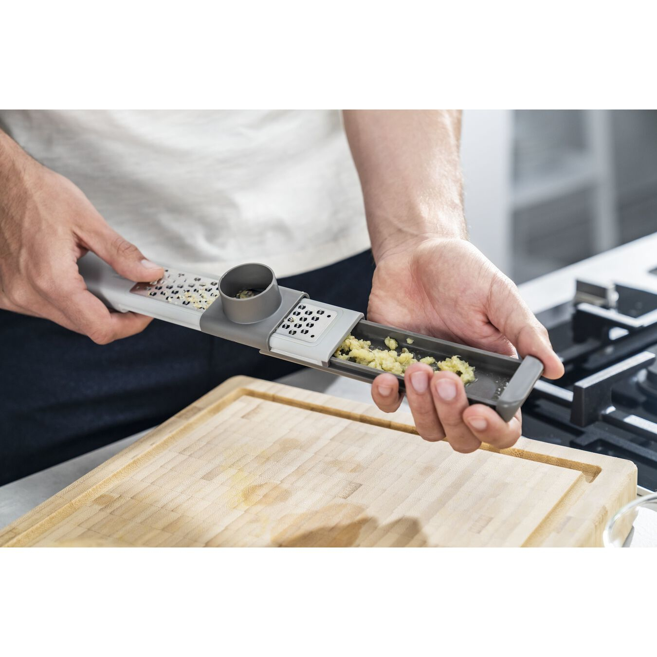 Person grating garlic with a gray and white kitchen tool on a wooden cutting board, a stove is in the background.