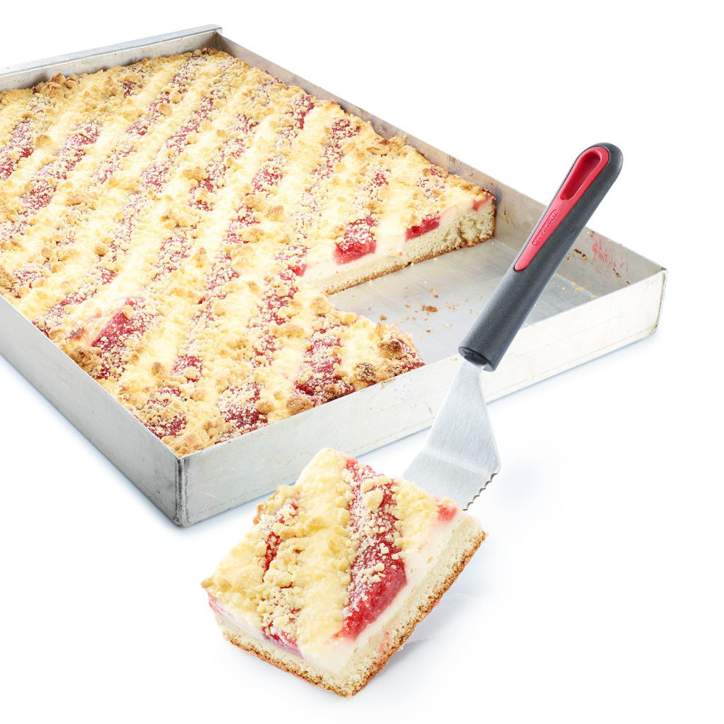 Ein quadratischer Kuchen mit einem herausgeschnittenen Stück auf einem Metalltablett. Ein Spatel hält ein Stück Kuchen.