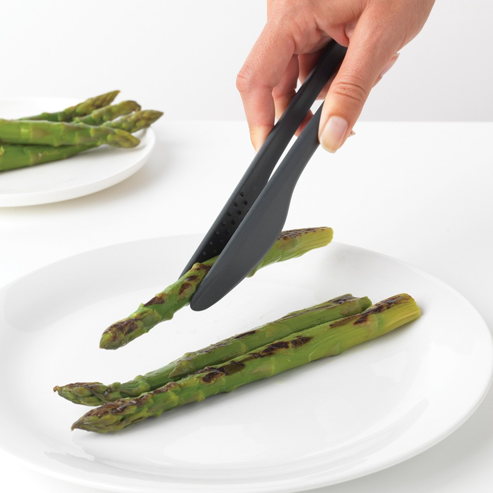 Mano che usa delle pinze per mettere gli asparagi grigliati su un piatto con altri asparagi.