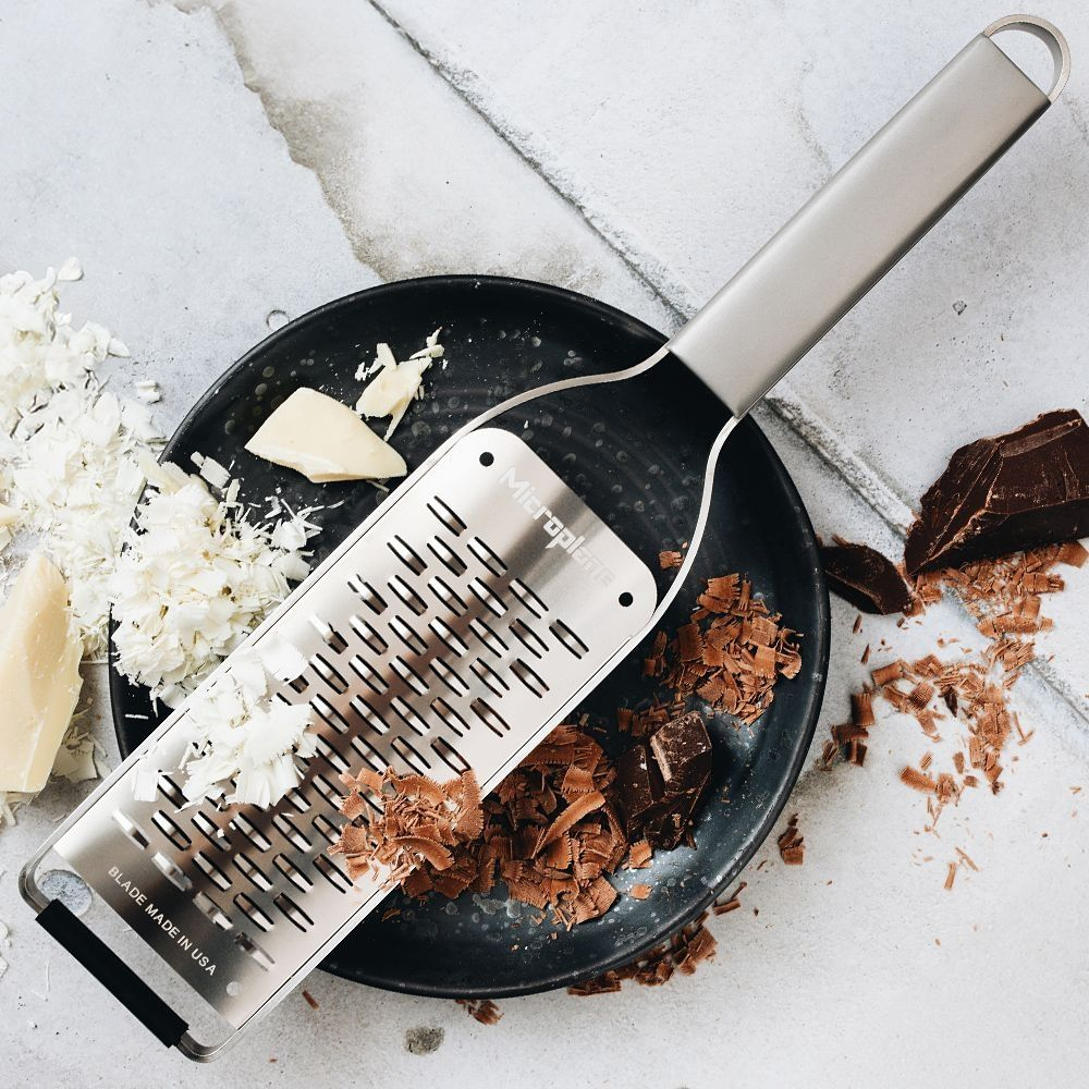 Grattugia Microplane su un piatto nero con formaggio, cioccolato e scaglie, su una superficie grigia.