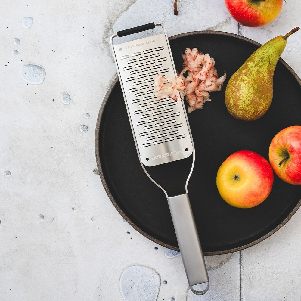 Grattugia Microplane su piatto nero con frutta grattugiata, pera e due mele su superficie grigia.