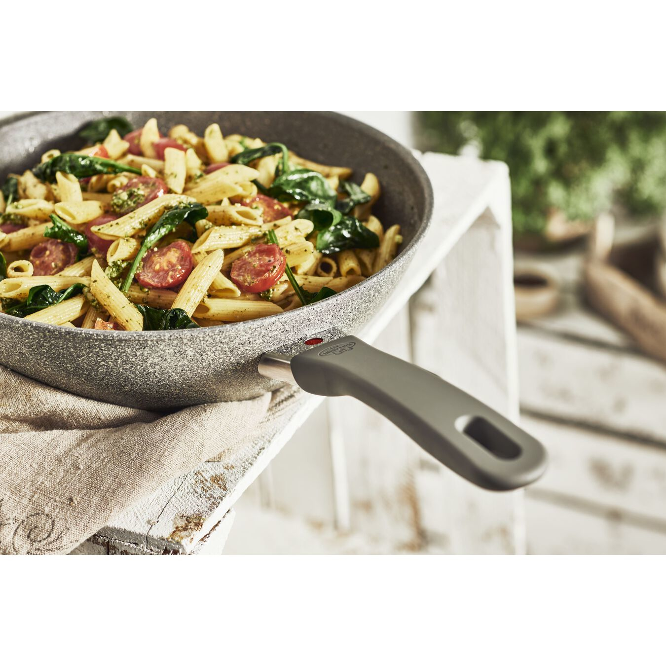 Nudeln mit Tomaten und Spinat in einer Pfanne. Graue Pfanne mit grauem Griff. Weißer Tisch.