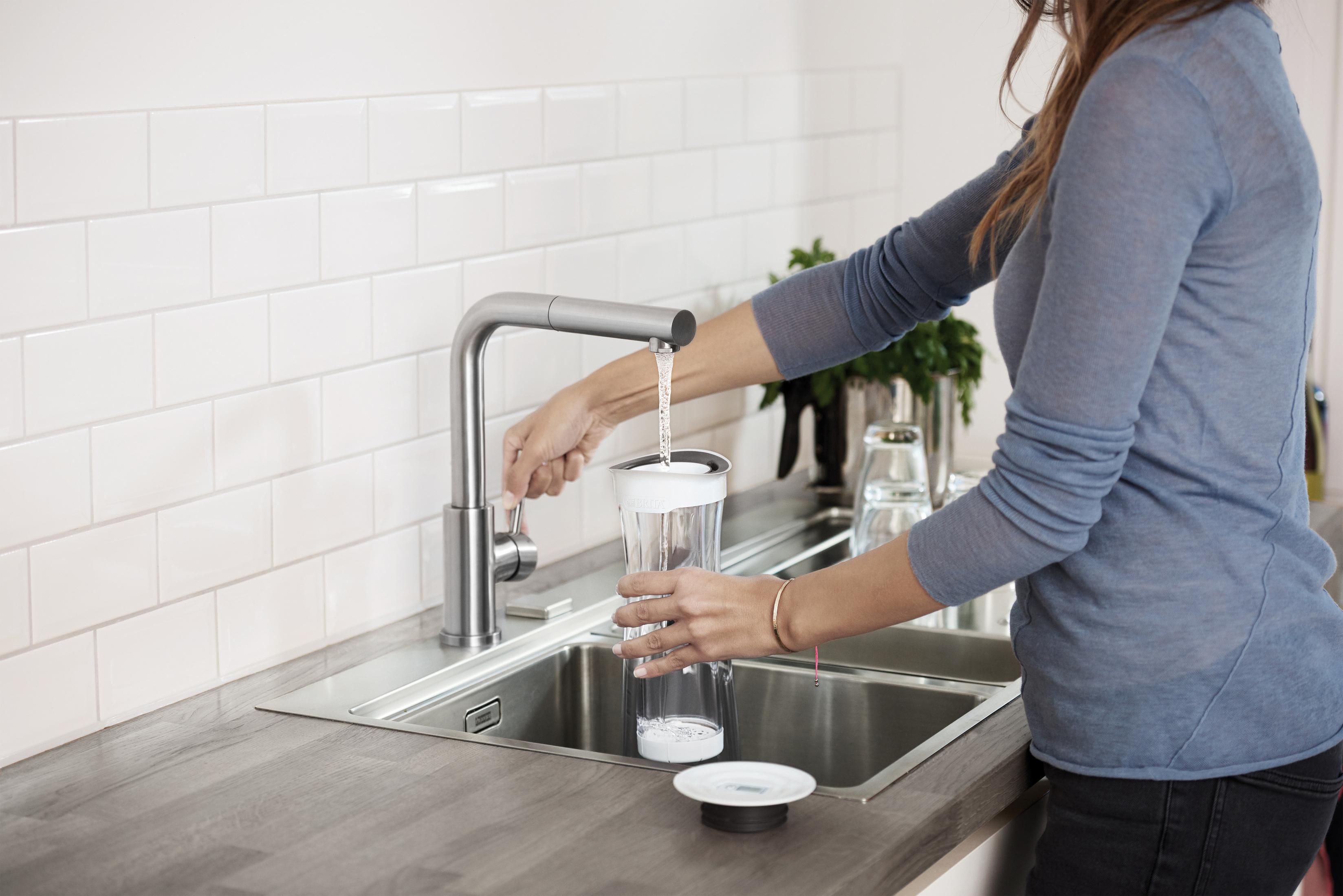 Eine Person, die eine klare Karaffe an einem Küchenbecken mit Wasser füllt.