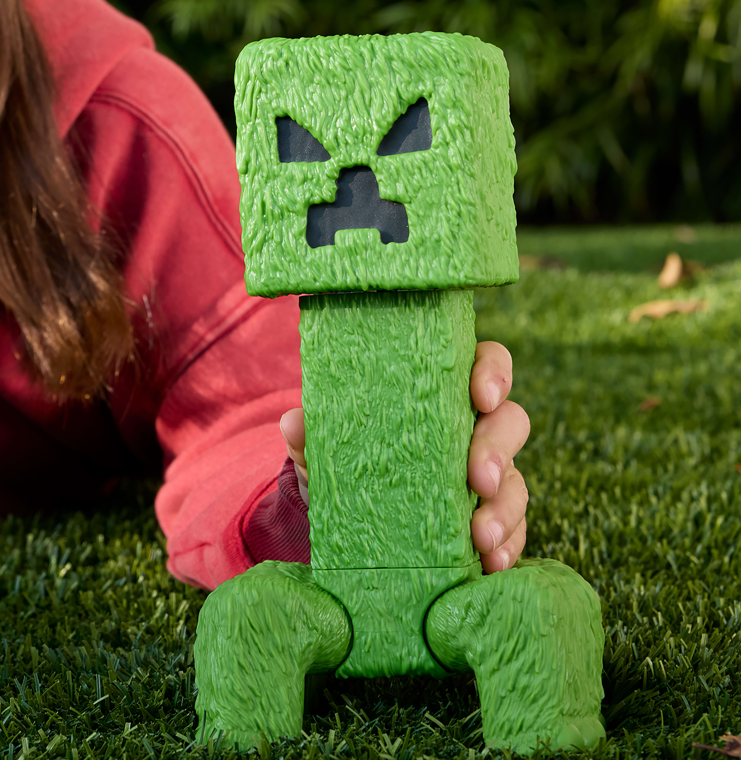 Ręka trzymająca zieloną zabawkę Minecraft Creeper na trawie, z czerwoną koszulą w tle.