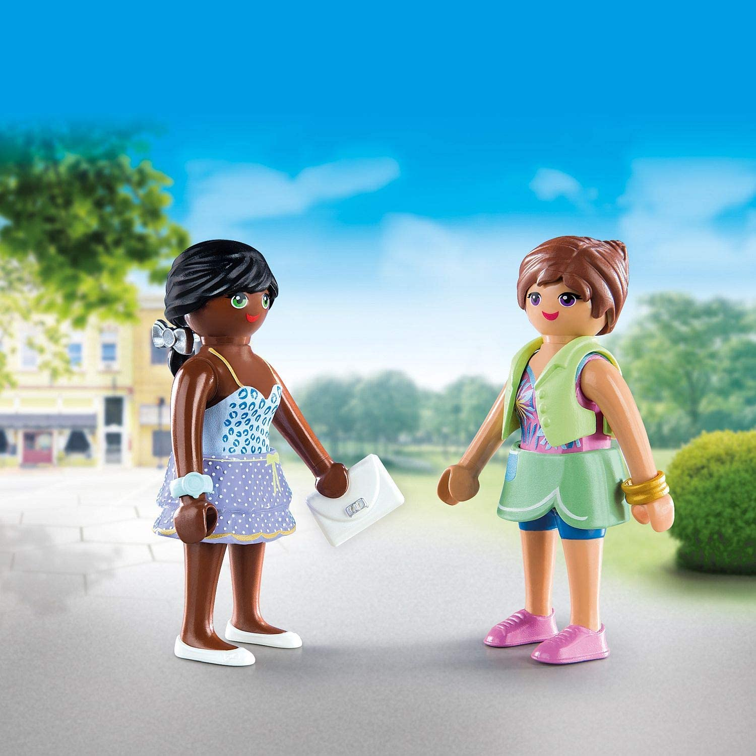 Zwei Playmobil-Figuren, eine hält einen Brief. Blauer Himmel, grünes Gras und Gebäude im Hintergrund.