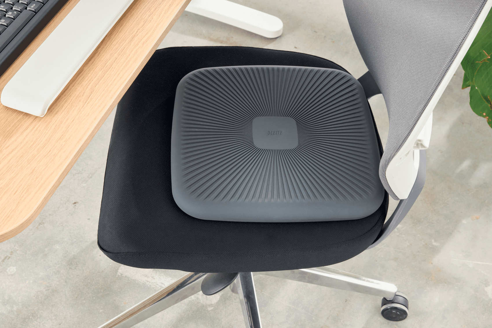 Szara poduszka na czarnym krześle biurowym, z biurkiem i częścią klawiatury w tle.