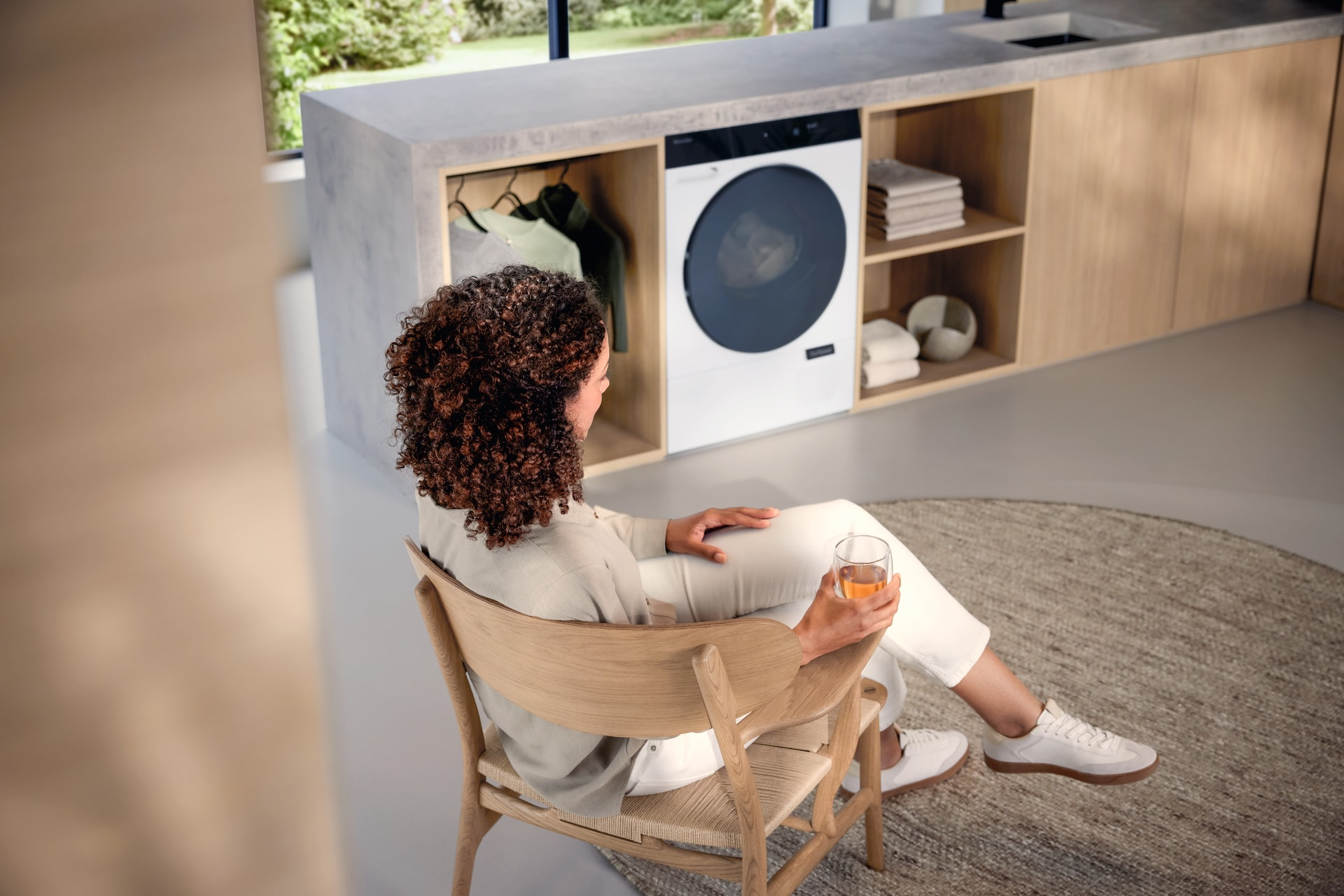 Donna seduta su una sedia di legno, tiene un bicchiere, in una lavanderia moderna. Lavatrice bianca.