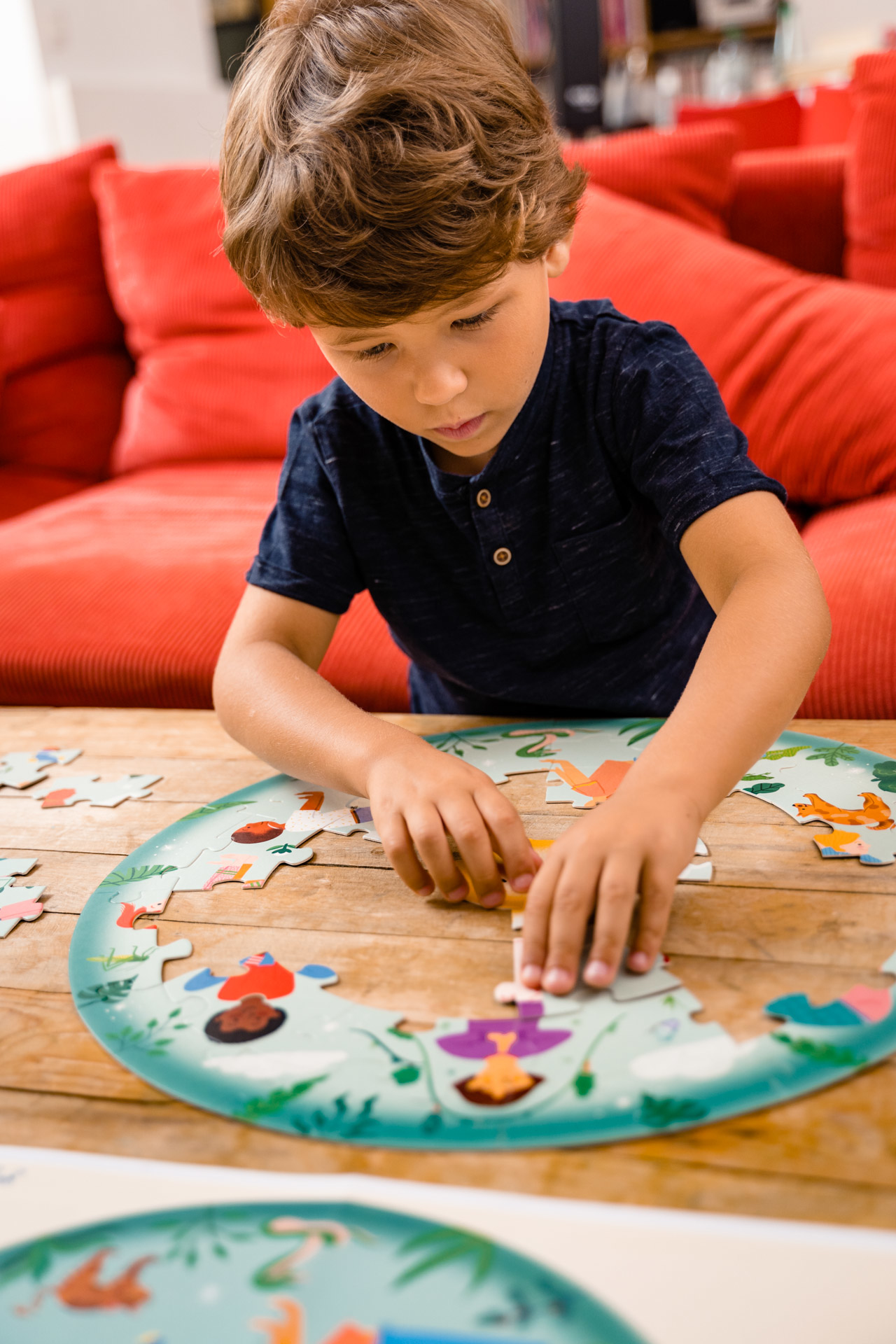 Ein Kind setzt ein rundes Puzzle auf einem Holztisch zusammen. Ein rotes Sofa ist im Hintergrund.
