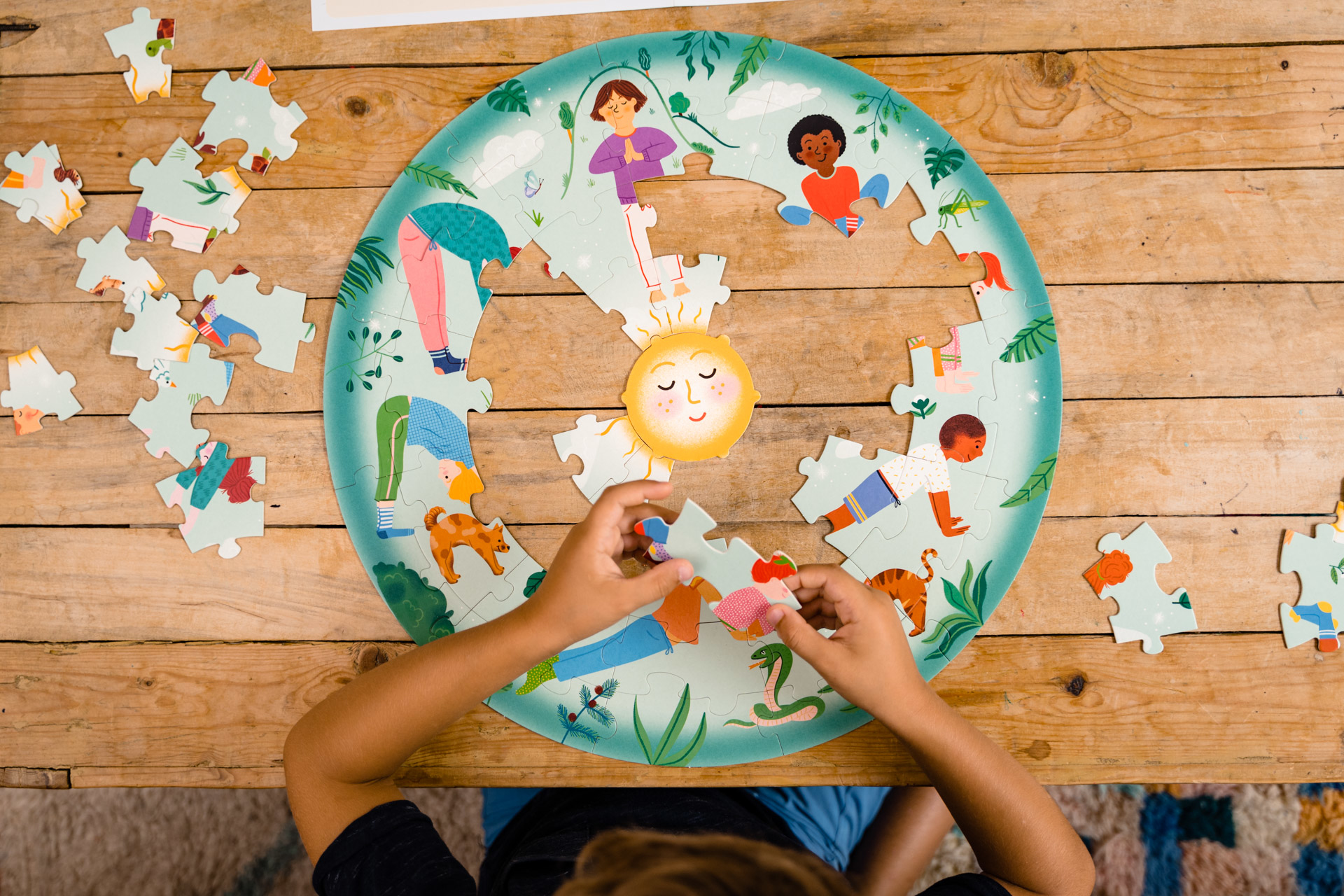 Kind, das ein Yoga-Puzzle auf einem Holztisch zusammensetzt. Figuren machen Yoga um eine Sonne herum. Andere Puzzleteile sind verstreut.