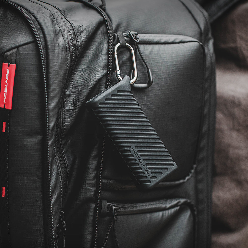 Czarny futerał przymocowany do plecaka za pomocą karabińczyka. Futerał jest prostokątny z żebrami.