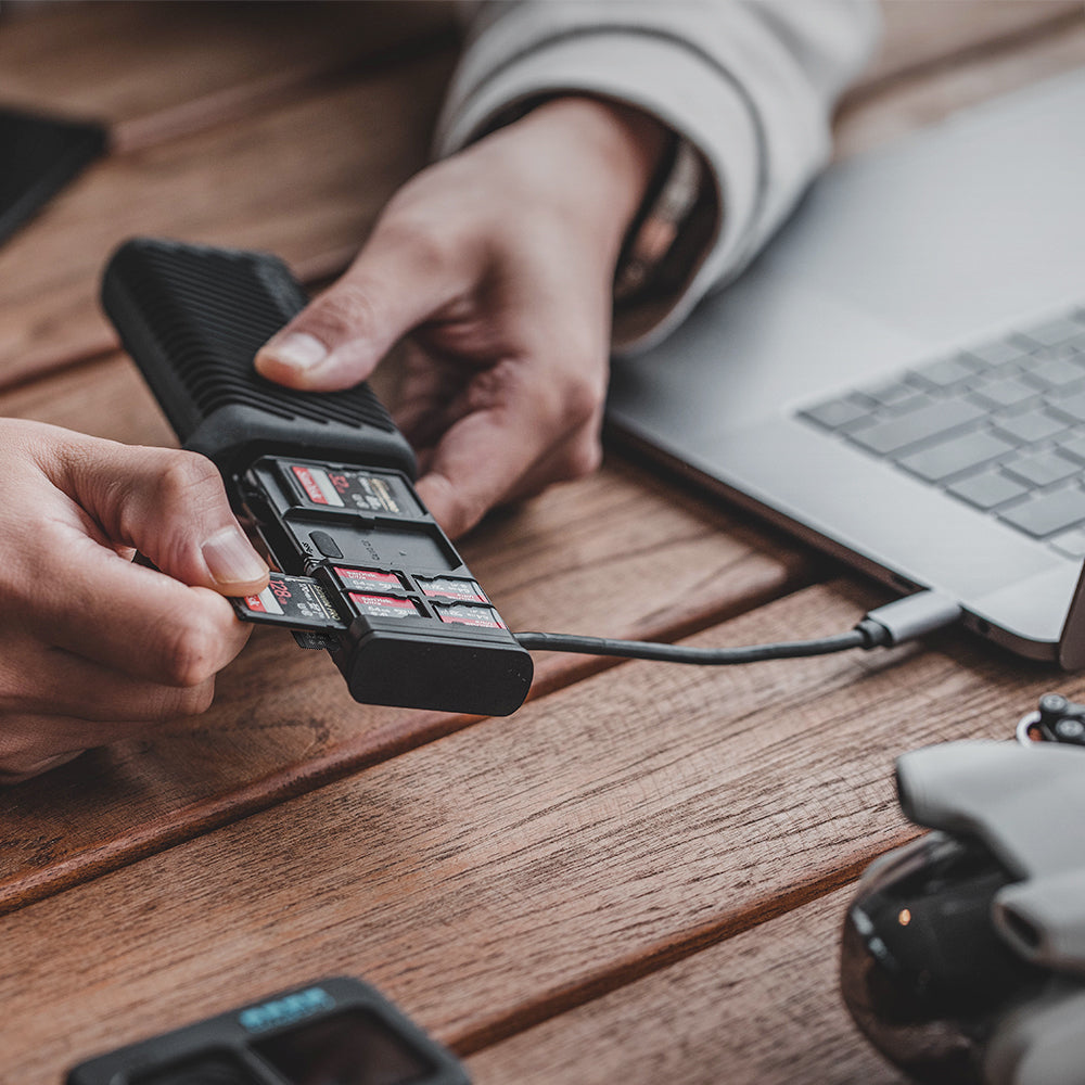 Ręce wkładające karty SD do czarnego czytnika kart, podłączonego do laptopa przez USB.