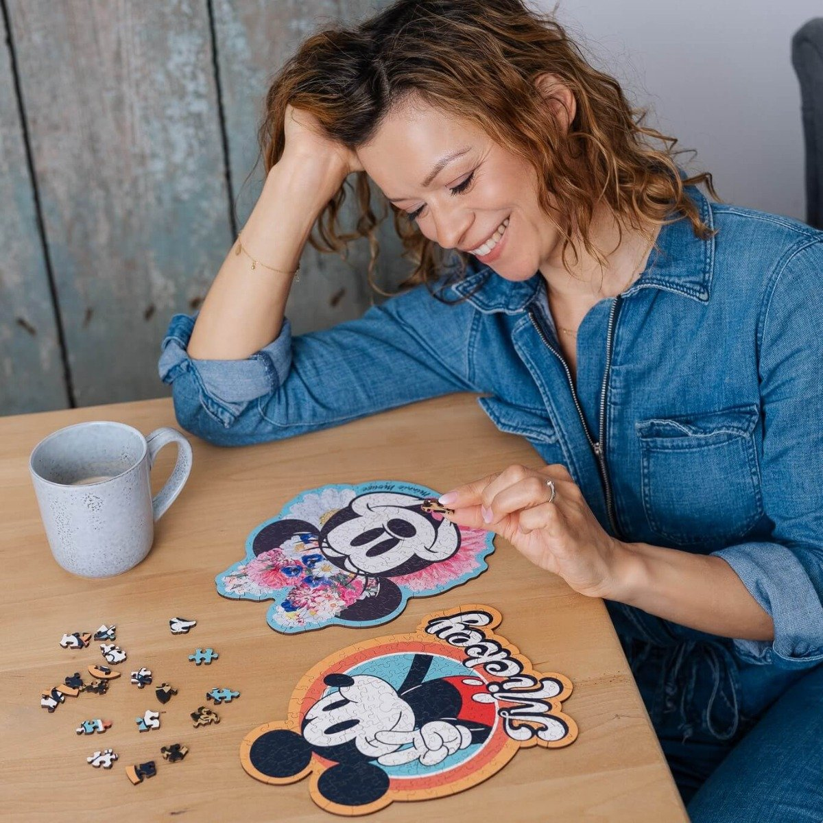 A woman is sitting at a table with two Minnie Mouse puzzles, smiling. There are puzzle pieces on the table.
